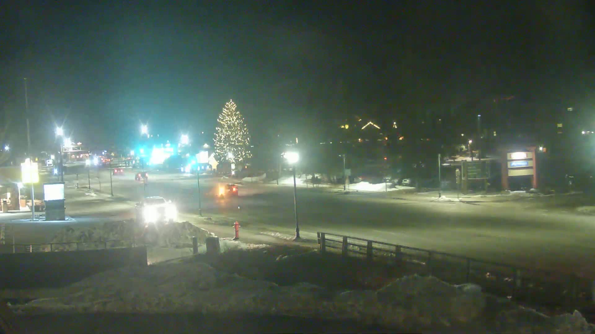 A snow-covered street at night, lit by streetlights, shows a few vehicles and a decorated Christmas tree in the center.