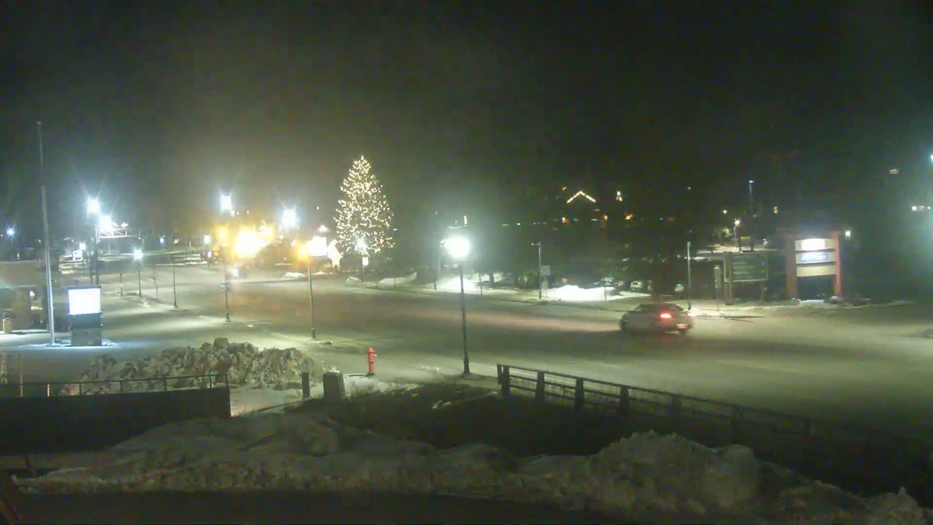 A snow-covered street at night, illuminated by streetlights, shows a single car driving past a decorated Christmas tree.