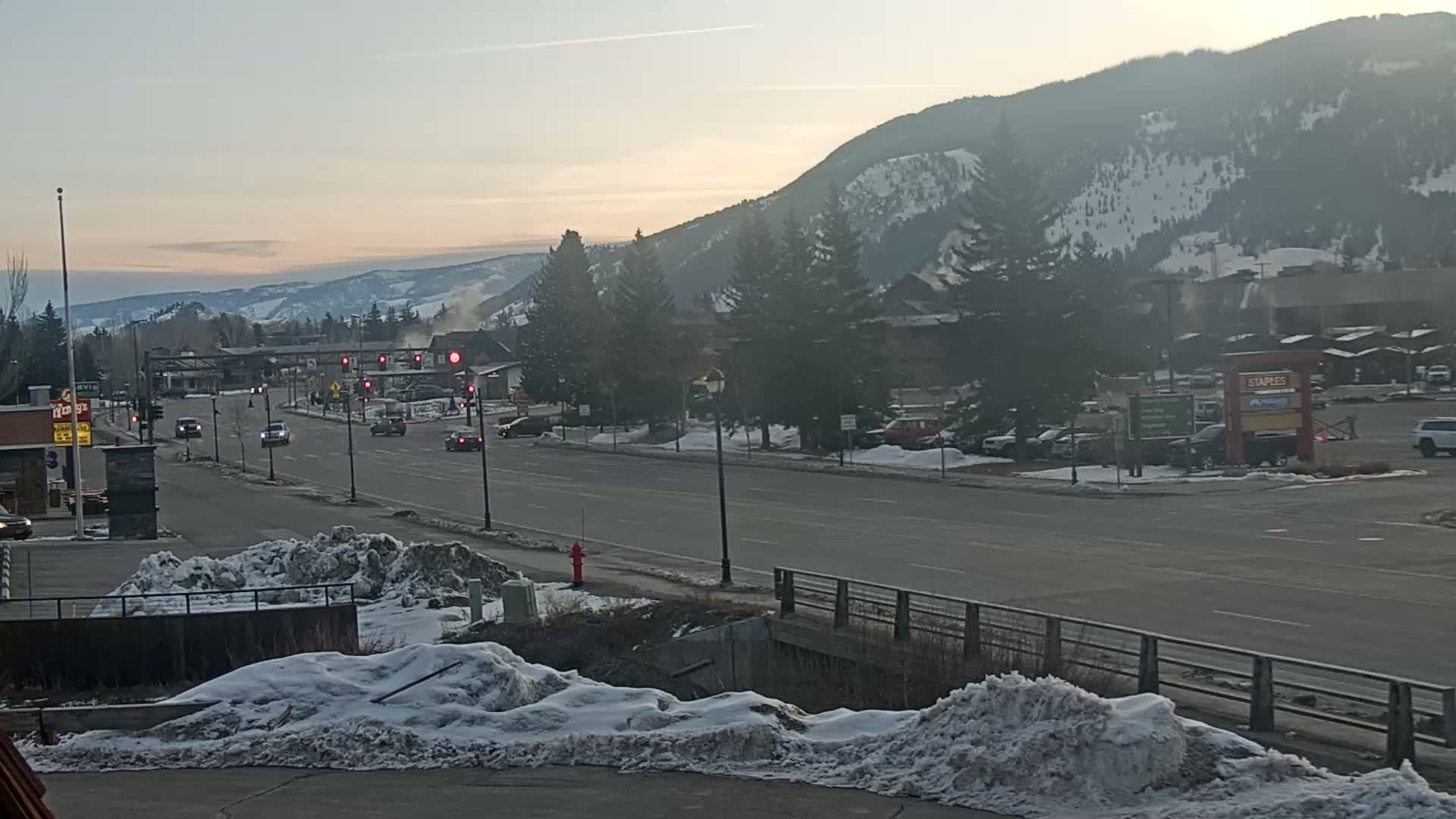 A snow-covered street scene shows a road with several cars,  lined with buildings and snow-dusted evergreen trees, in front of snow-capped mountains under a dusky sky.