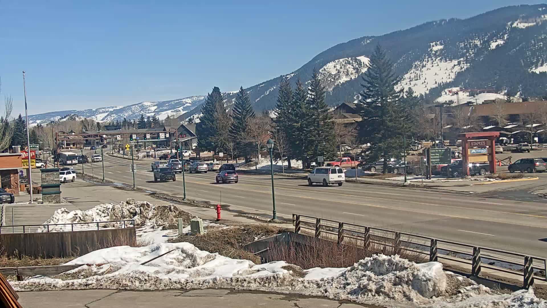 A sunny day reveals a town street with cars driving past buildings and snow-covered patches on the sides.  Snow-capped mountains are visible in the background.