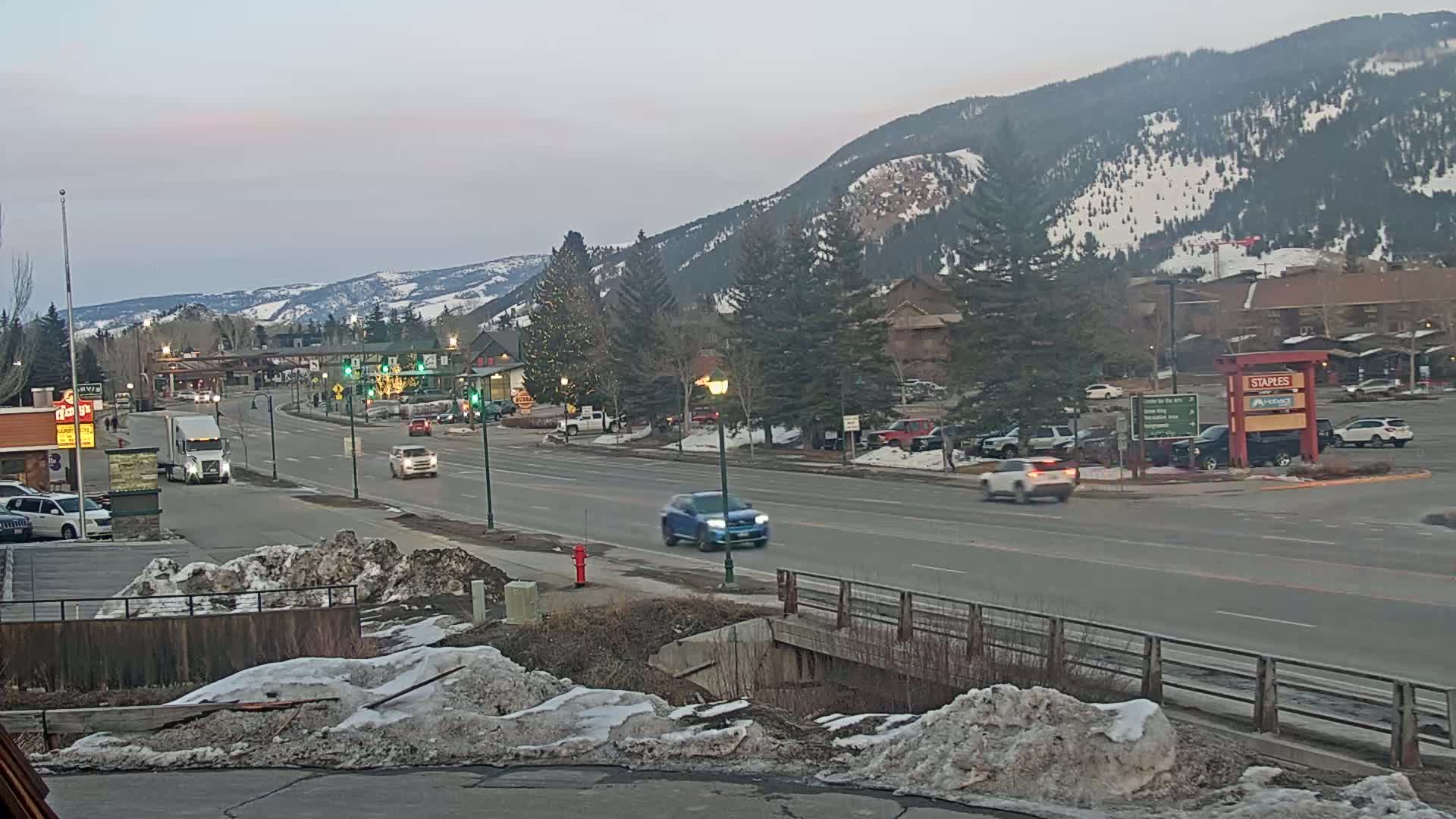 A twilight view of a snow-dusted mountain town, with several cars driving on a multi-lane road that passes by businesses and residential areas.