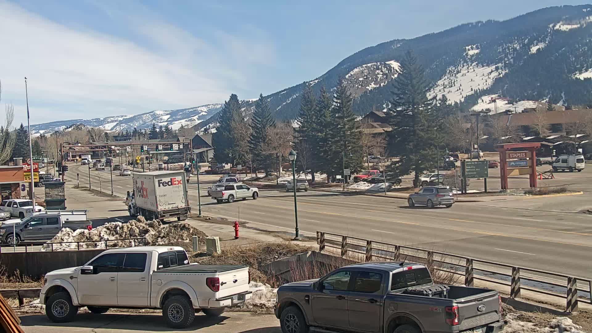 A sunny day reveals a mountain town street scene with snow-capped mountains in the background, several vehicles driving on the road, and parked trucks in the foreground.