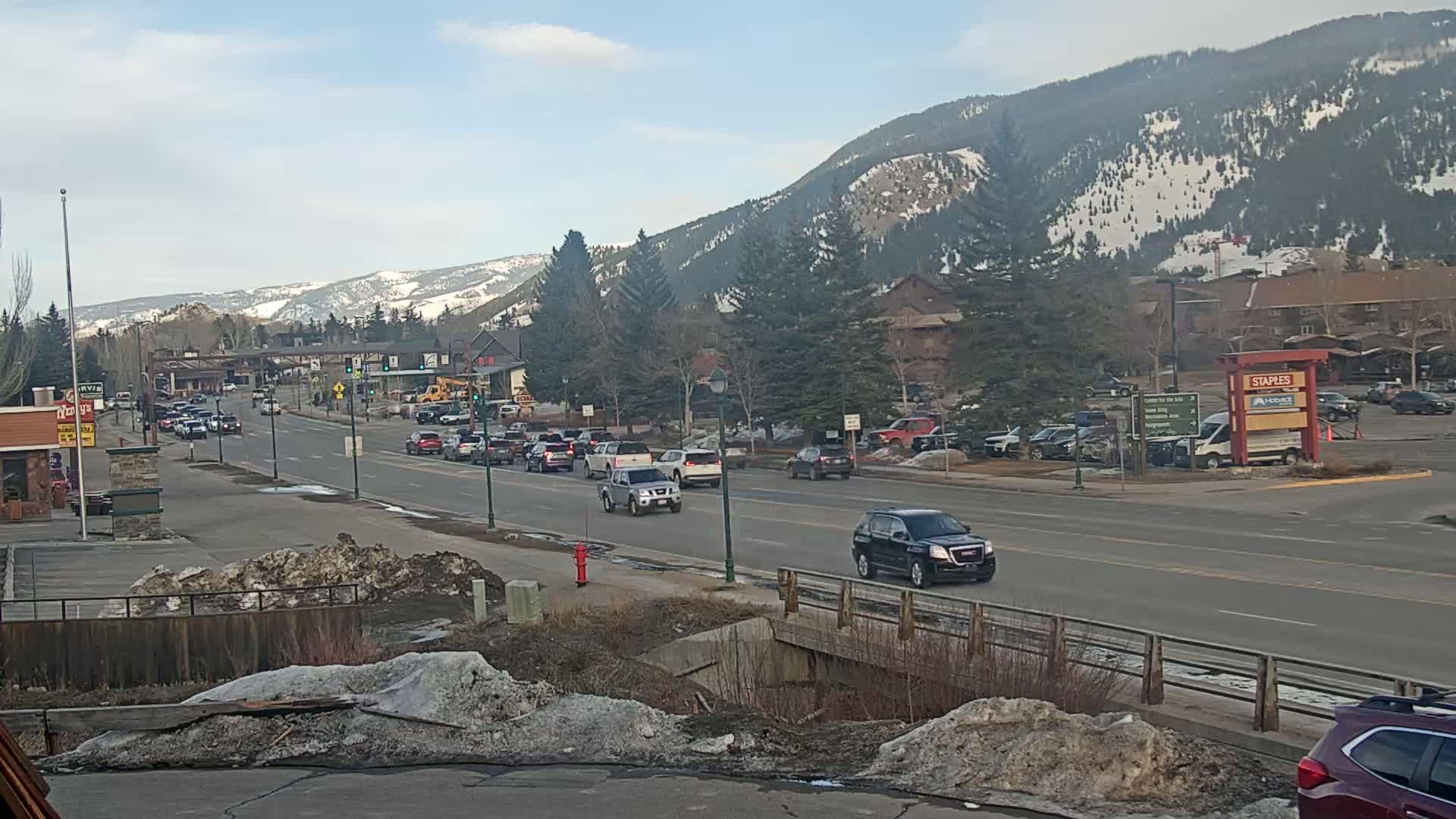 A sunny day in a mountain town shows moderate traffic on a street lined with buildings and trees, with snow-capped mountains in the background.