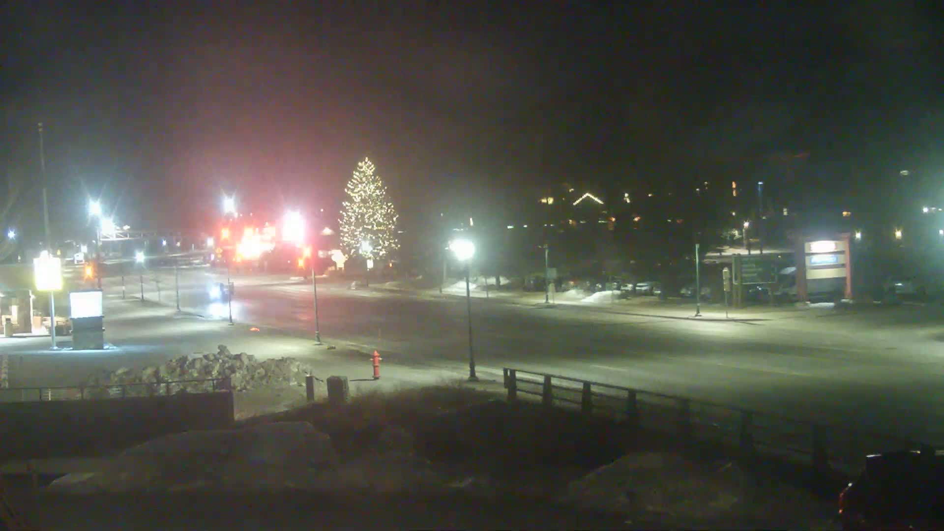 A snow-covered street at night, illuminated by streetlights, with a decorated Christmas tree visible in the mid-ground and buildings in the background.