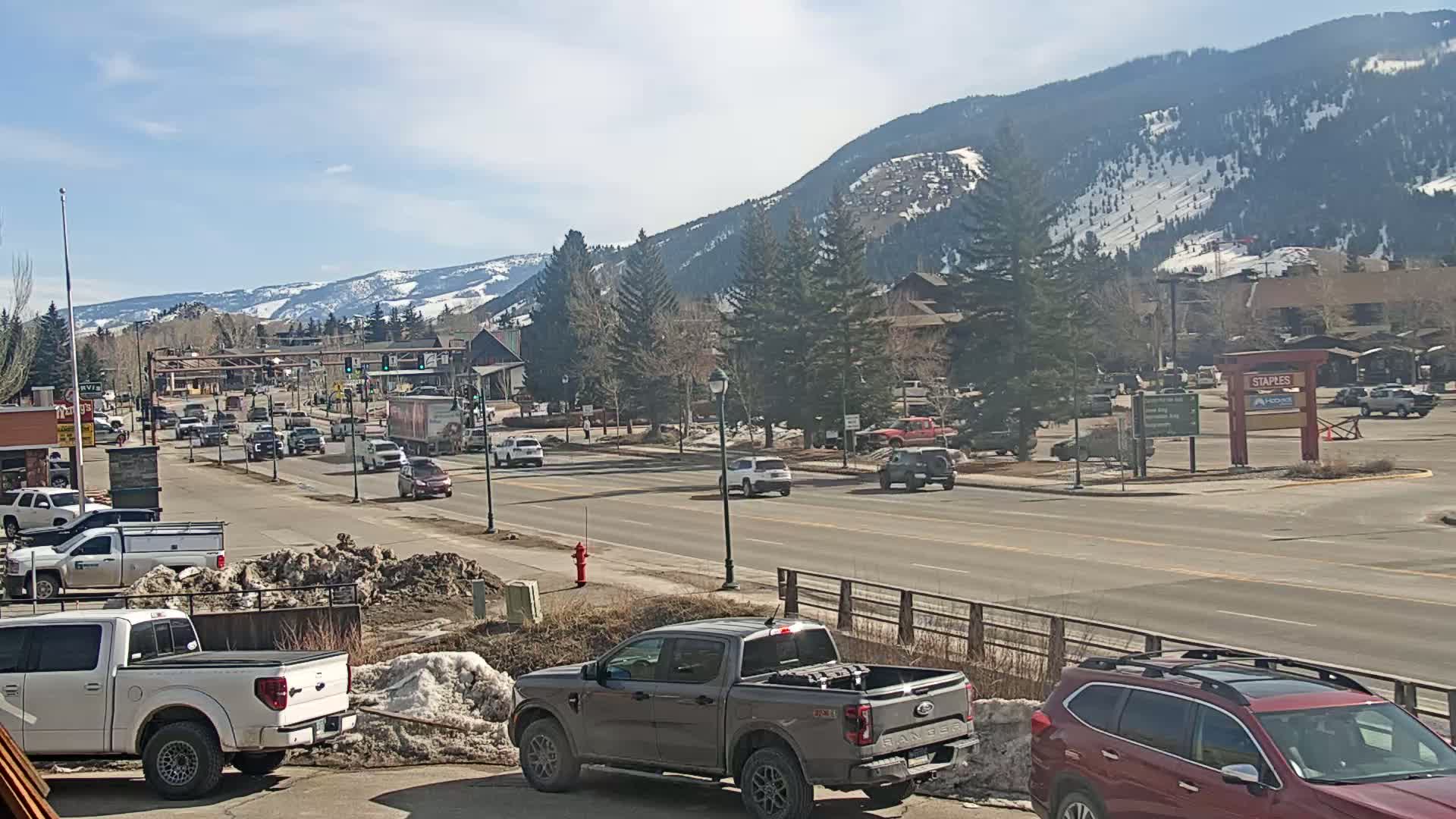 A sunny day reveals a road scene with moderate traffic, snow-capped mountains in the background, and patchy snow on the ground near the road.