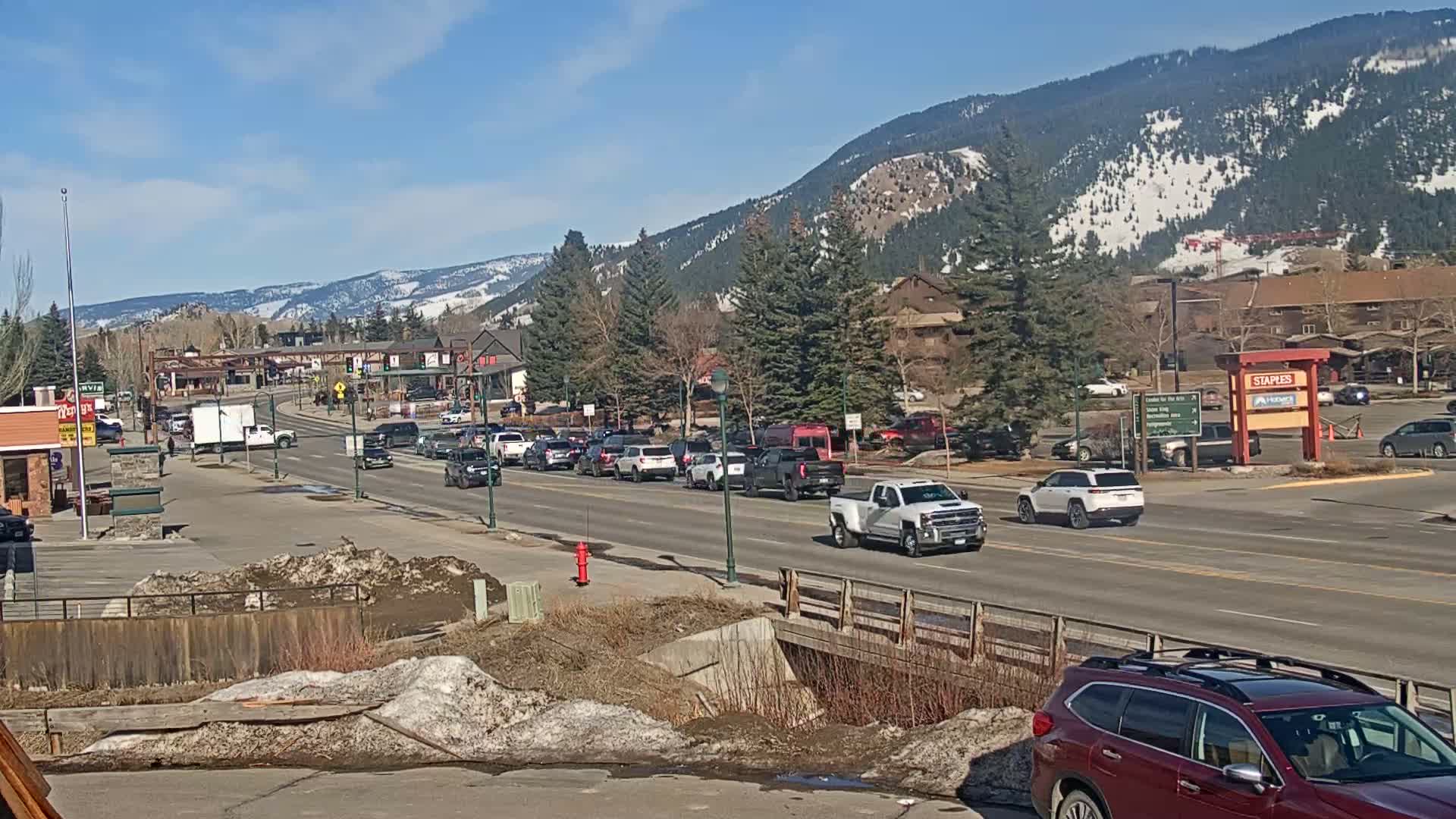 A sunny day reveals a mountain town with a busy street featuring several cars and light traffic, and snow-dusted mountains in the background.