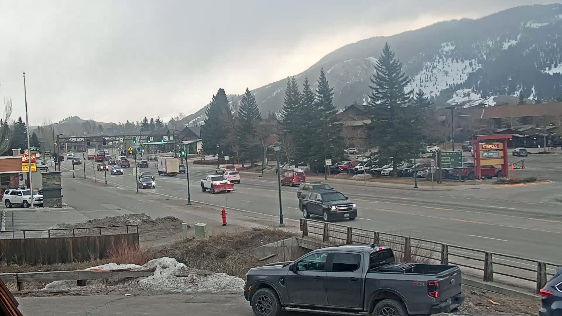A multi-lane road with moderate traffic flows through a town, under an overcast sky, with snow-capped mountains in the background.