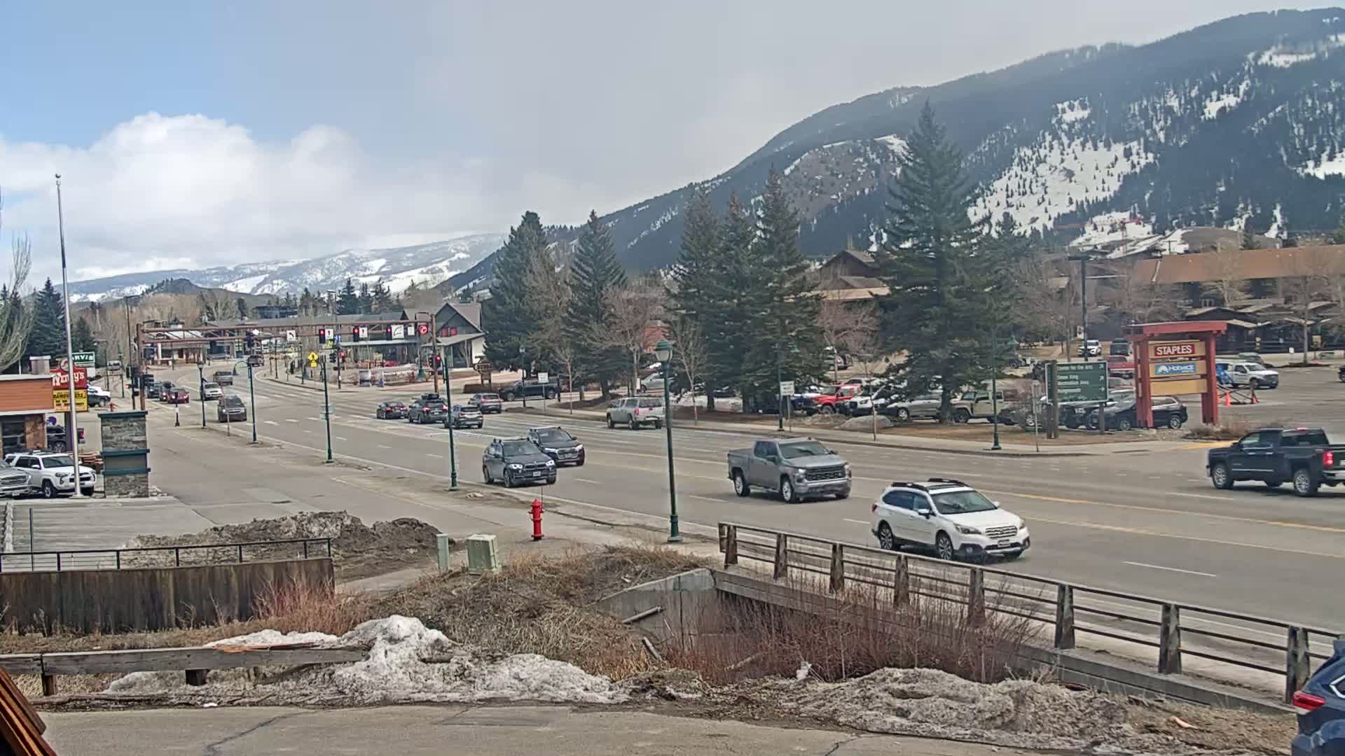 A partly cloudy day reveals a mountain-lined town with a multi-lane road featuring several cars, some parked and some driving, and a small bridge in the foreground.