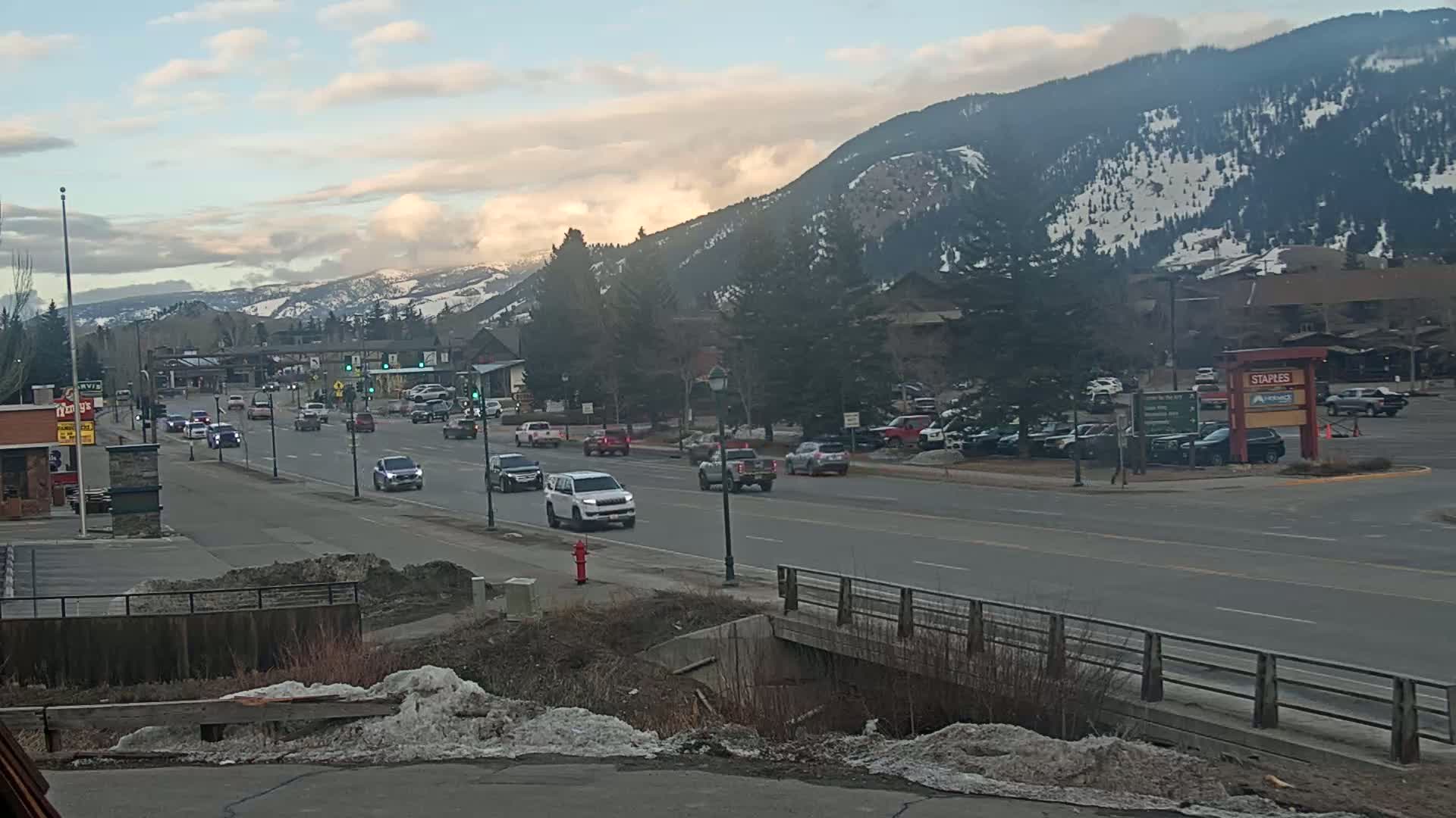 A partly cloudy day shows a road with moderate traffic and some snow on the ground, with mountains in the background showing some snow.