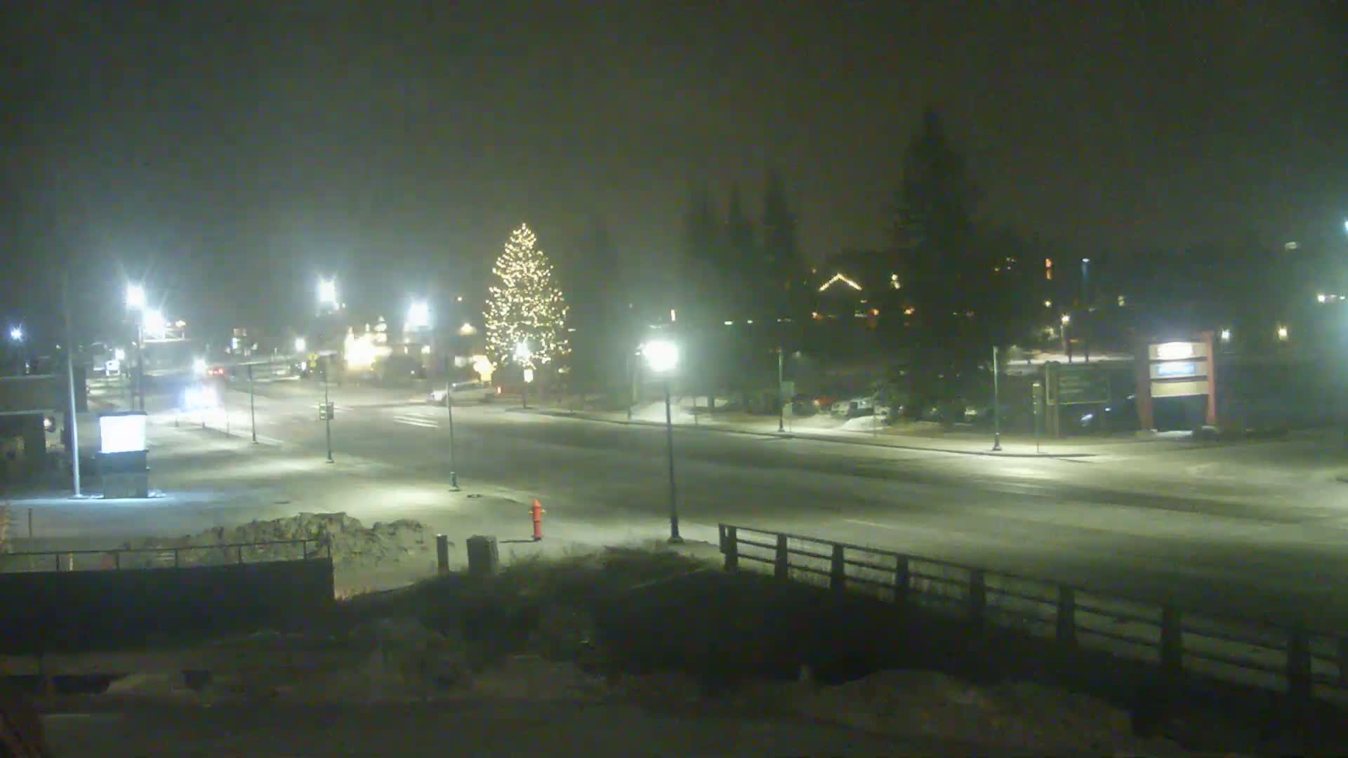A snow-covered road at night is lit by streetlights, with a decorated Christmas tree visible in the center and cars sparsely driving along the street.