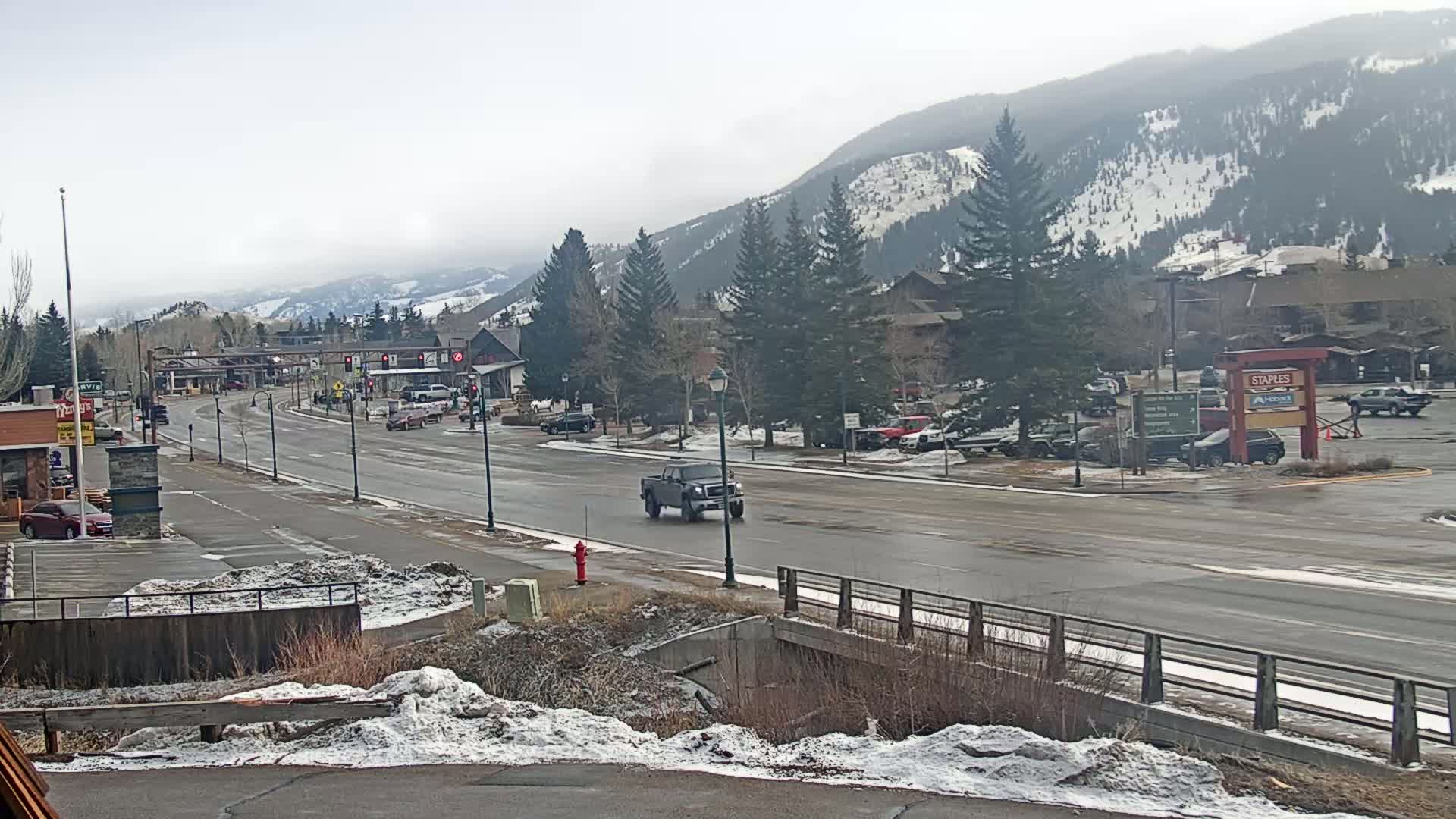 A wet, paved road stretches through a town nestled in a valley, with snow-capped mountains in the background and sparsely scattered snow on the ground.