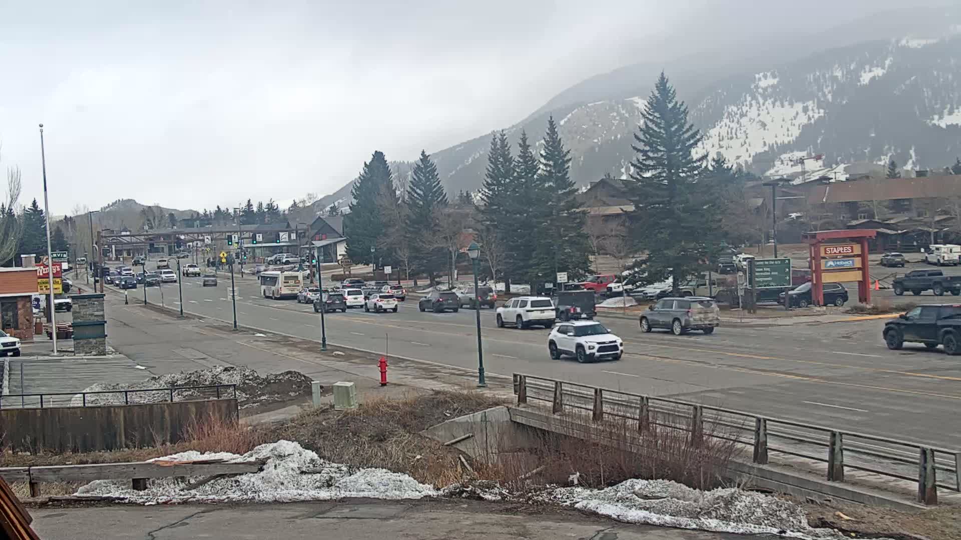 A partly cloudy day shows a multi-lane road with moderate traffic in a town nestled in a valley, with snow-dusted mountains in the background.