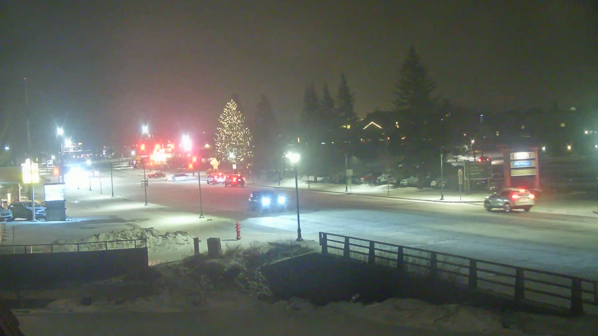 A snow-covered street at night, with several cars driving and a decorated Christmas tree visible in the background, under hazy conditions.