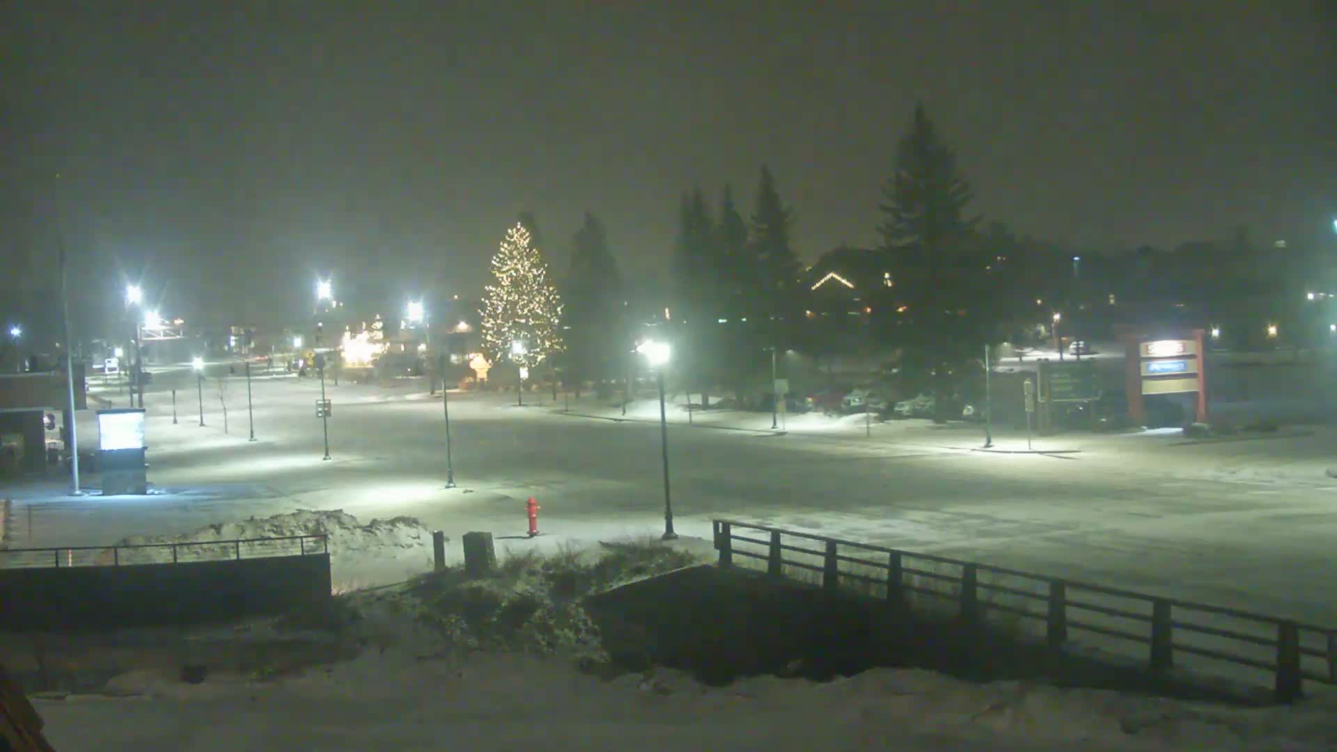 A snow-covered town square at night is illuminated by streetlights, with a decorated Christmas tree visible in the center.