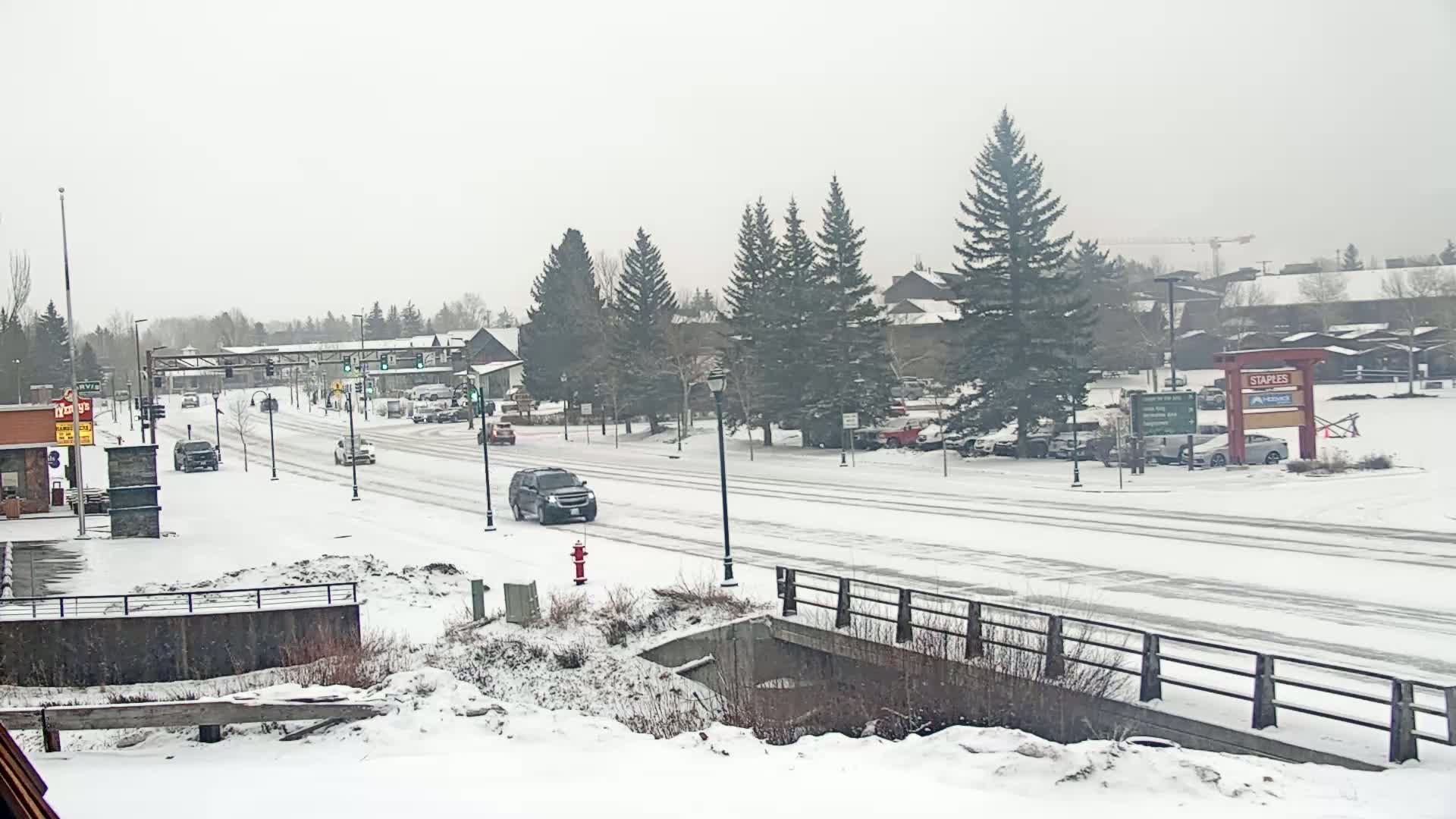A snow-covered street scene shows several vehicles driving on a main road lined with evergreen trees and buildings, under a light snowfall.