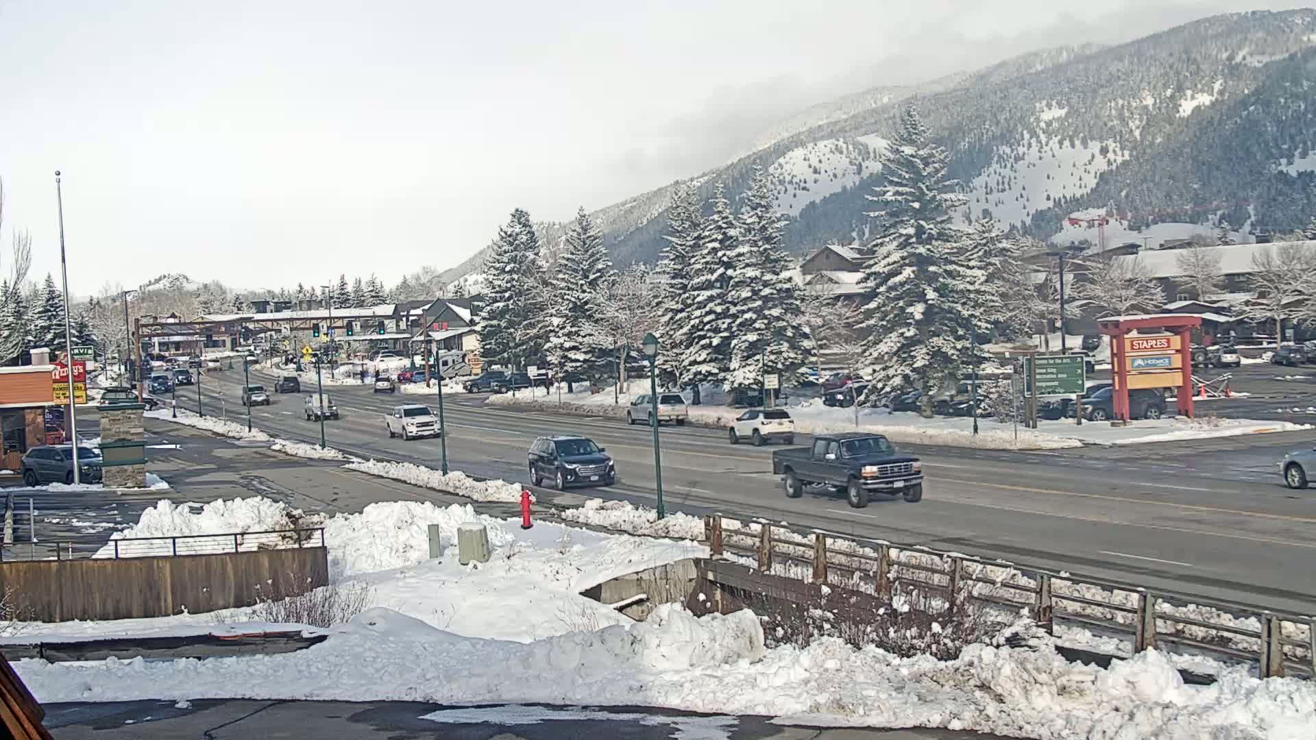 A snow-covered town with cars driving on a road, surrounded by snow-covered evergreens and mountains in the background under a bright, mostly clear sky.