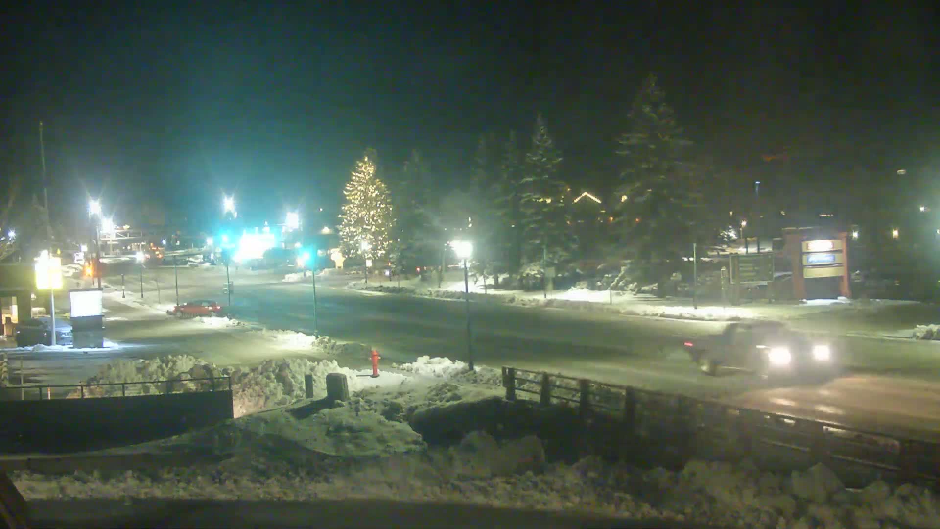 A snow-covered street at night, illuminated by streetlights, with a few vehicles and a decorated Christmas tree visible in the distance.