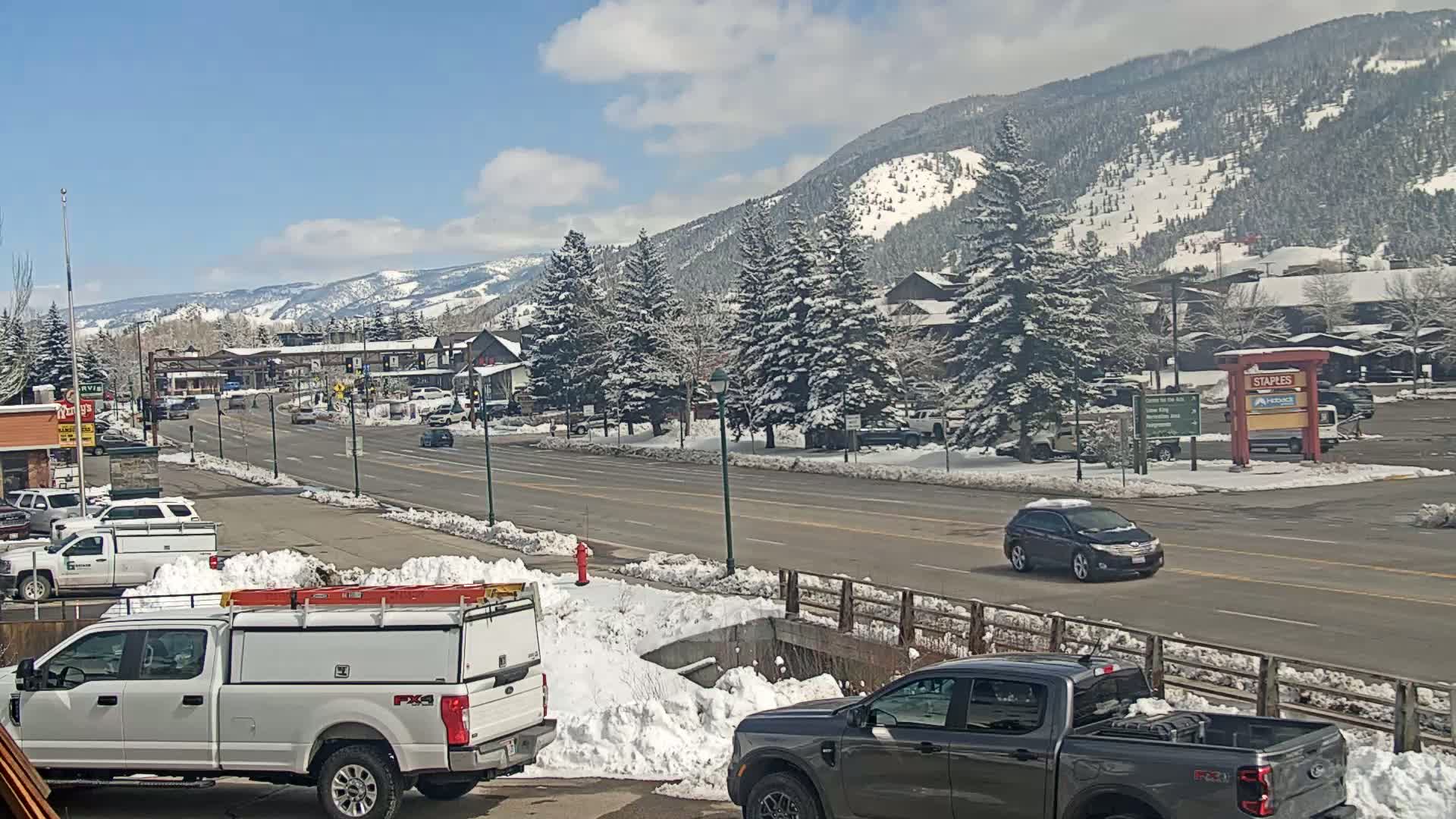 A snow-covered mountain town street scene on a sunny day, with several vehicles parked and driving, snowbanks lining the road, and snow-covered evergreen trees in the background.