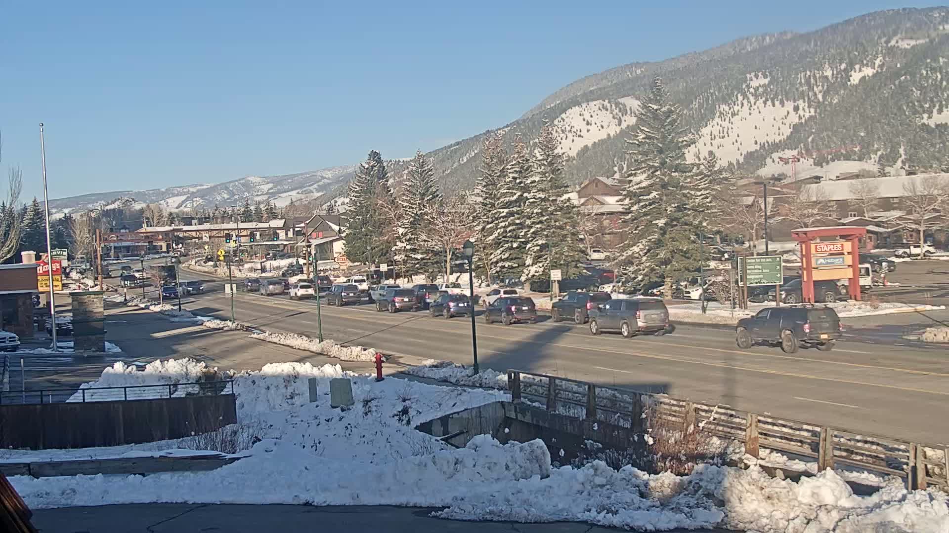 A line of cars drives along a road through a snow-covered town nestled in a sunny valley beneath snow-dusted mountains.