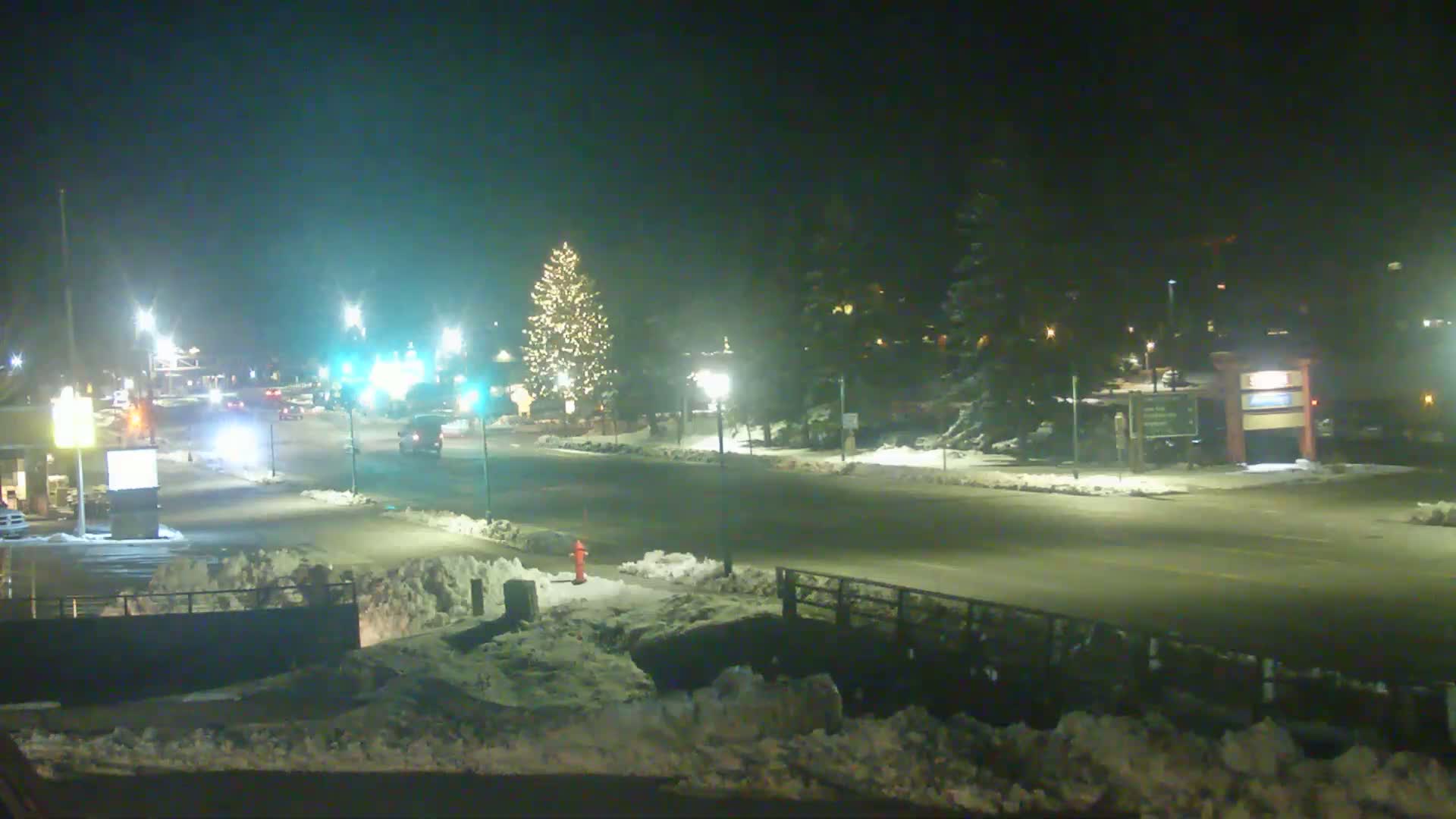 A snow-covered street at night is dimly lit by streetlights, with a few vehicles visible and a decorated Christmas tree in the background.