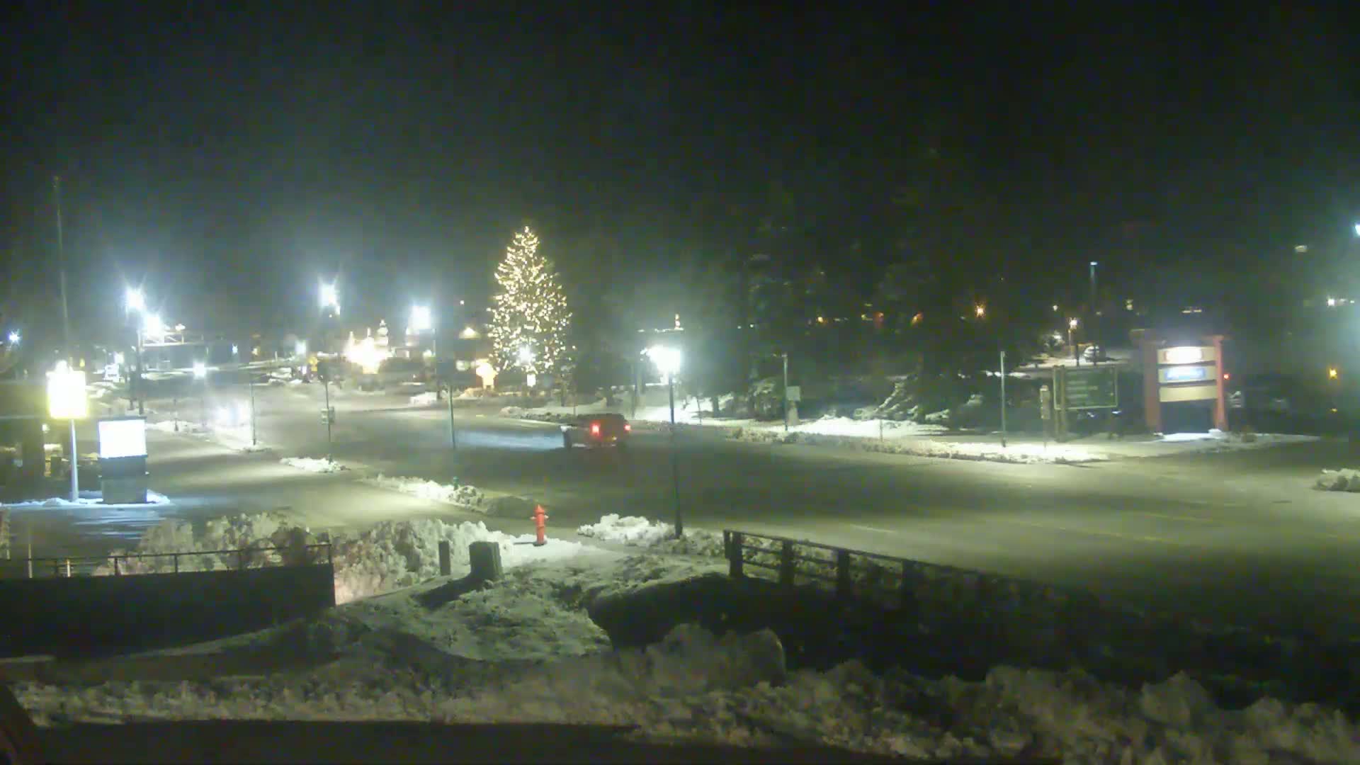 A snow-covered street at night, illuminated by streetlights, shows a single vehicle passing under a lit Christmas tree.