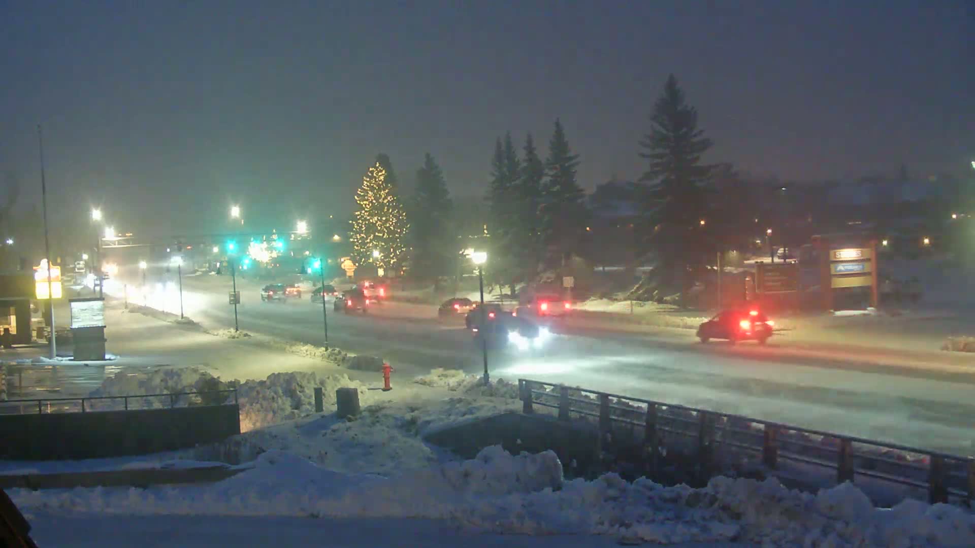 A snow-covered street at night, with cars driving through falling snow and a decorated Christmas tree visible in the background.