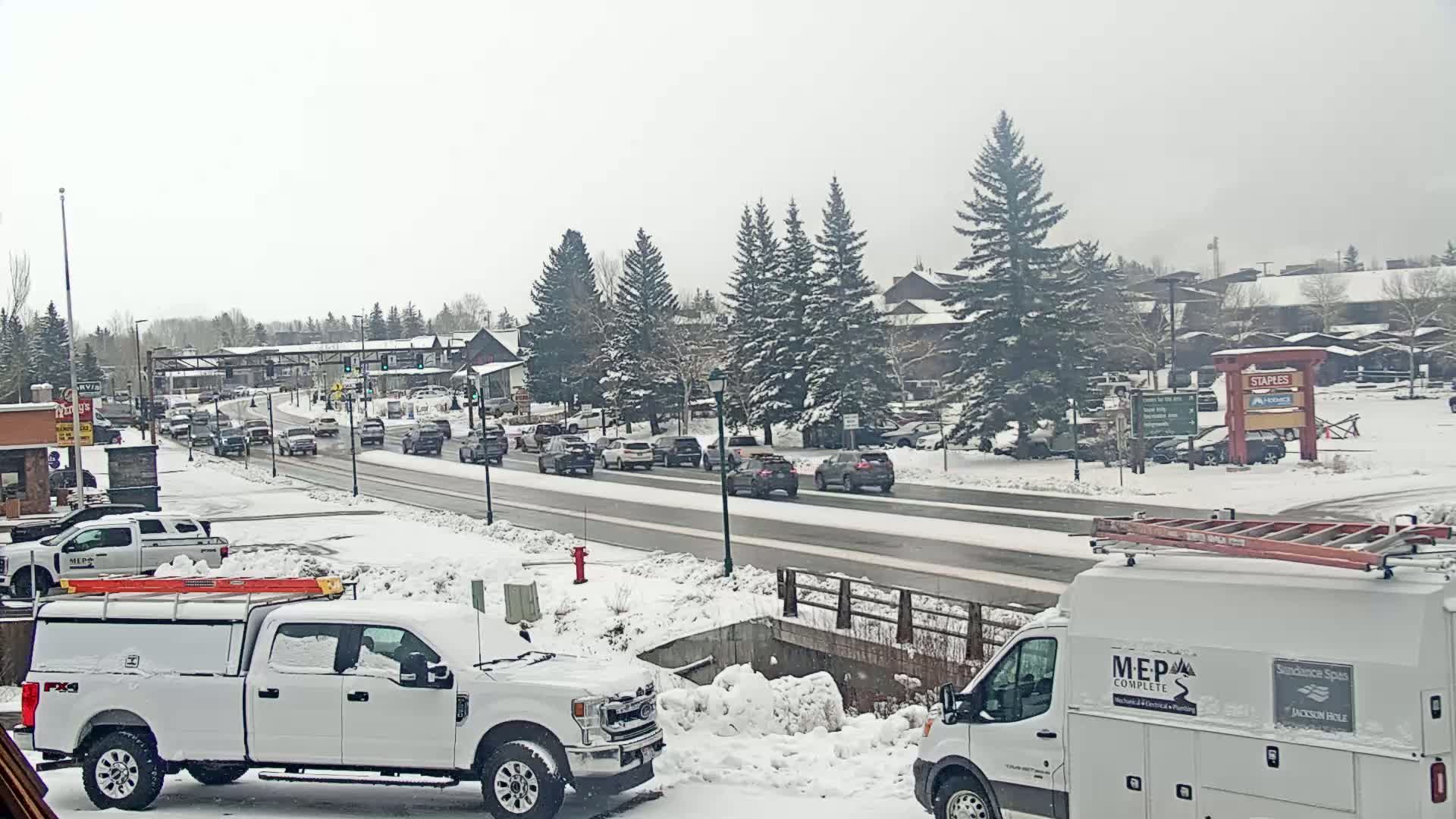 A snow-covered street scene shows a line of cars and trucks, including two service vehicles, parked along a road bordered by snow-covered evergreen trees and buildings.