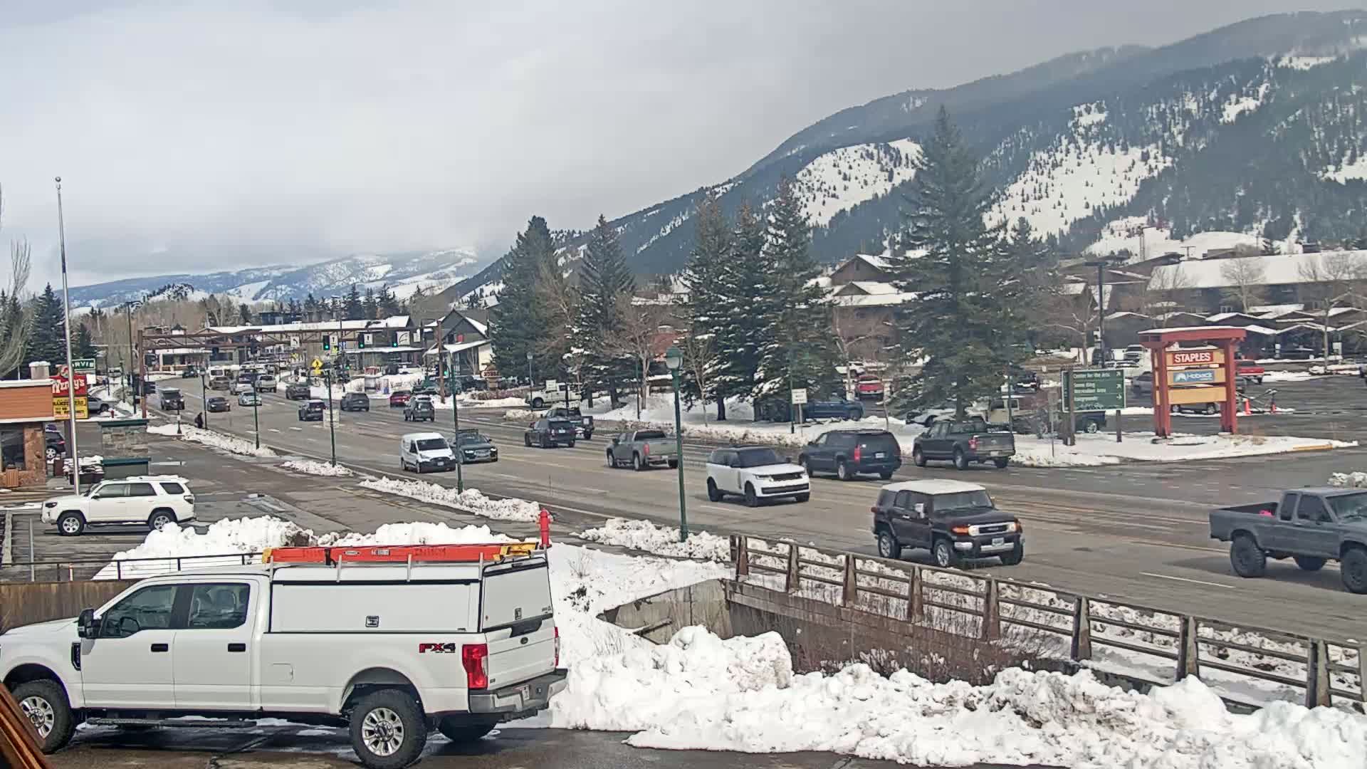 A snow-covered town with moderate traffic on a main road, nestled against a mountain range under an overcast sky.