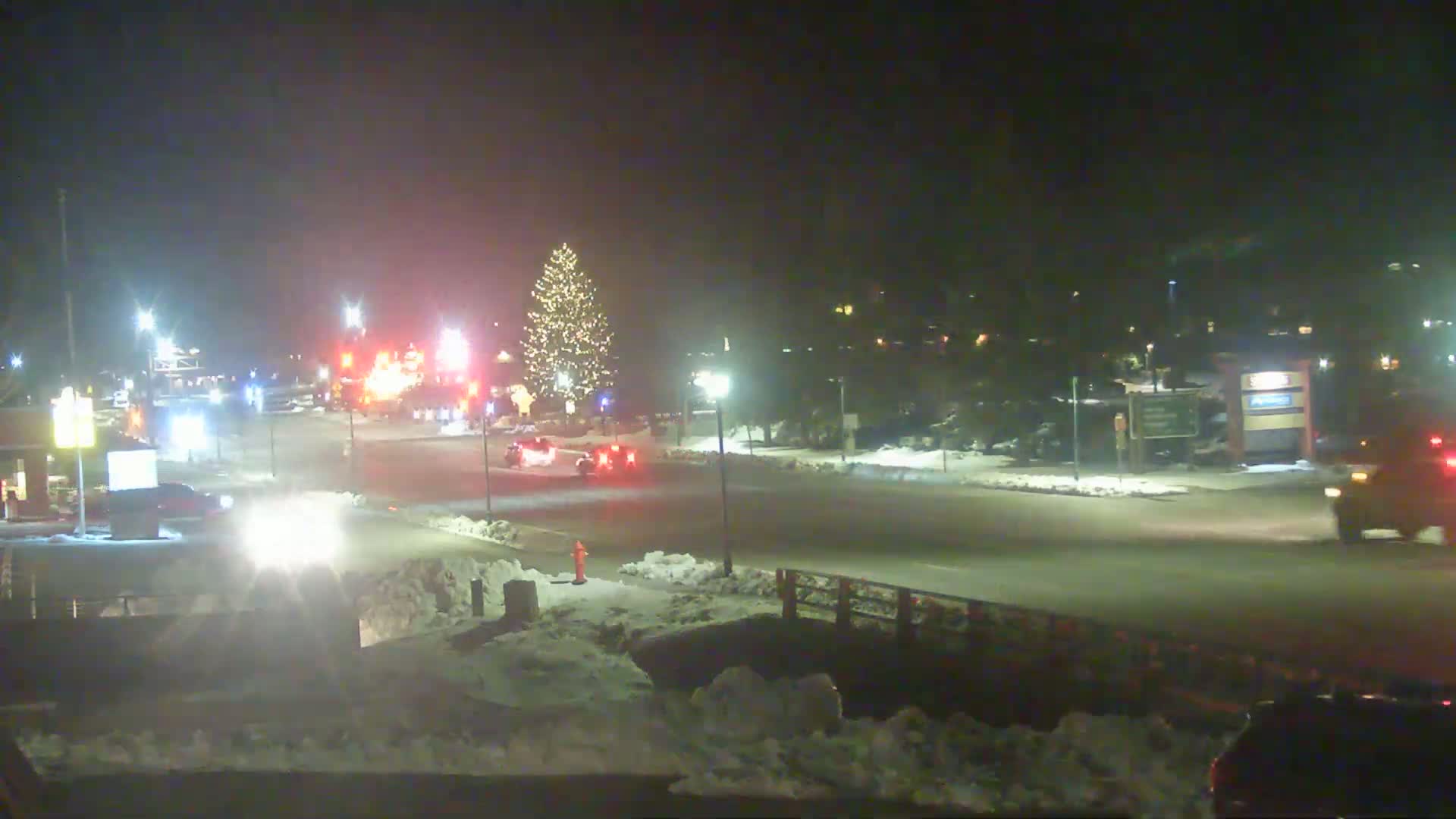 A nighttime street scene shows a snow-covered intersection with several vehicles and a decorated Christmas tree, under lightly snowy conditions.