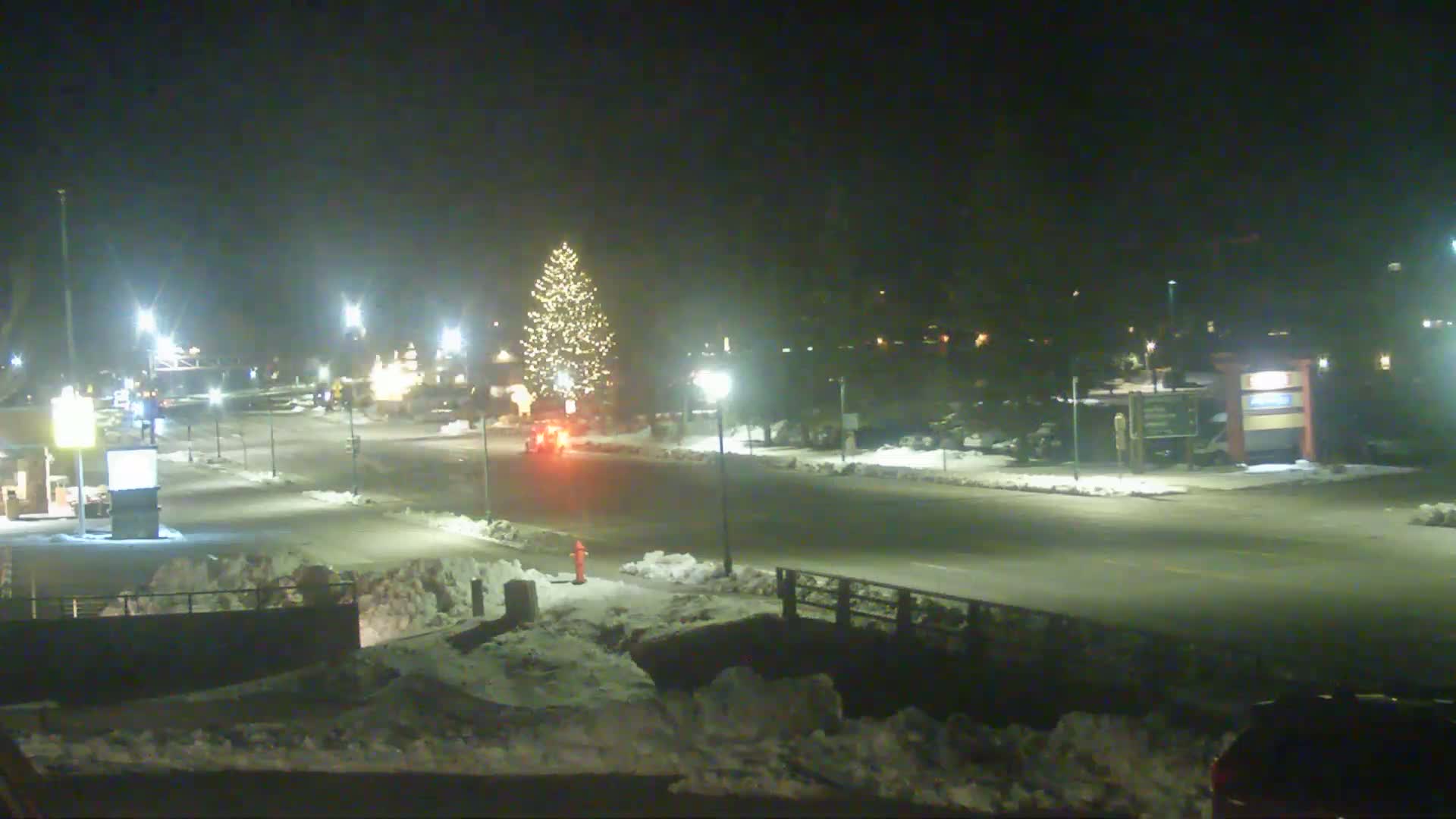A snow-covered street at night is lit by streetlights, featuring a decorated Christmas tree in the center and a few vehicles on the road.