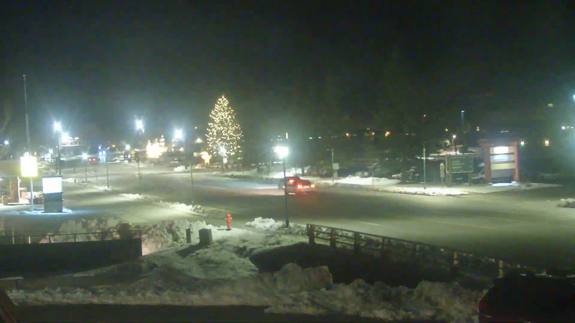 A snow-covered street scene at night shows a decorated Christmas tree and a vehicle driving past streetlights.