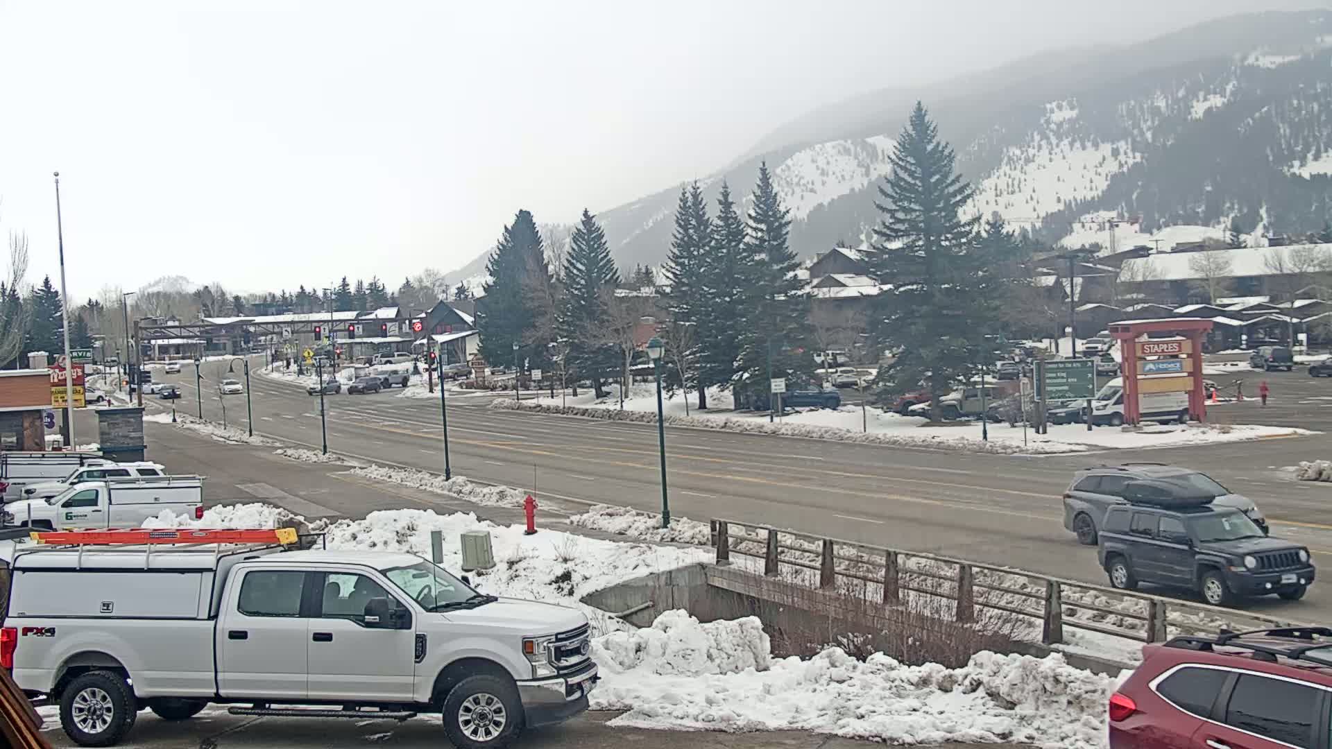 A snow-covered town nestled in a mountain valley under an overcast sky features a multi-lane road with several vehicles, including a white pickup truck, and a parking lot with various cars.