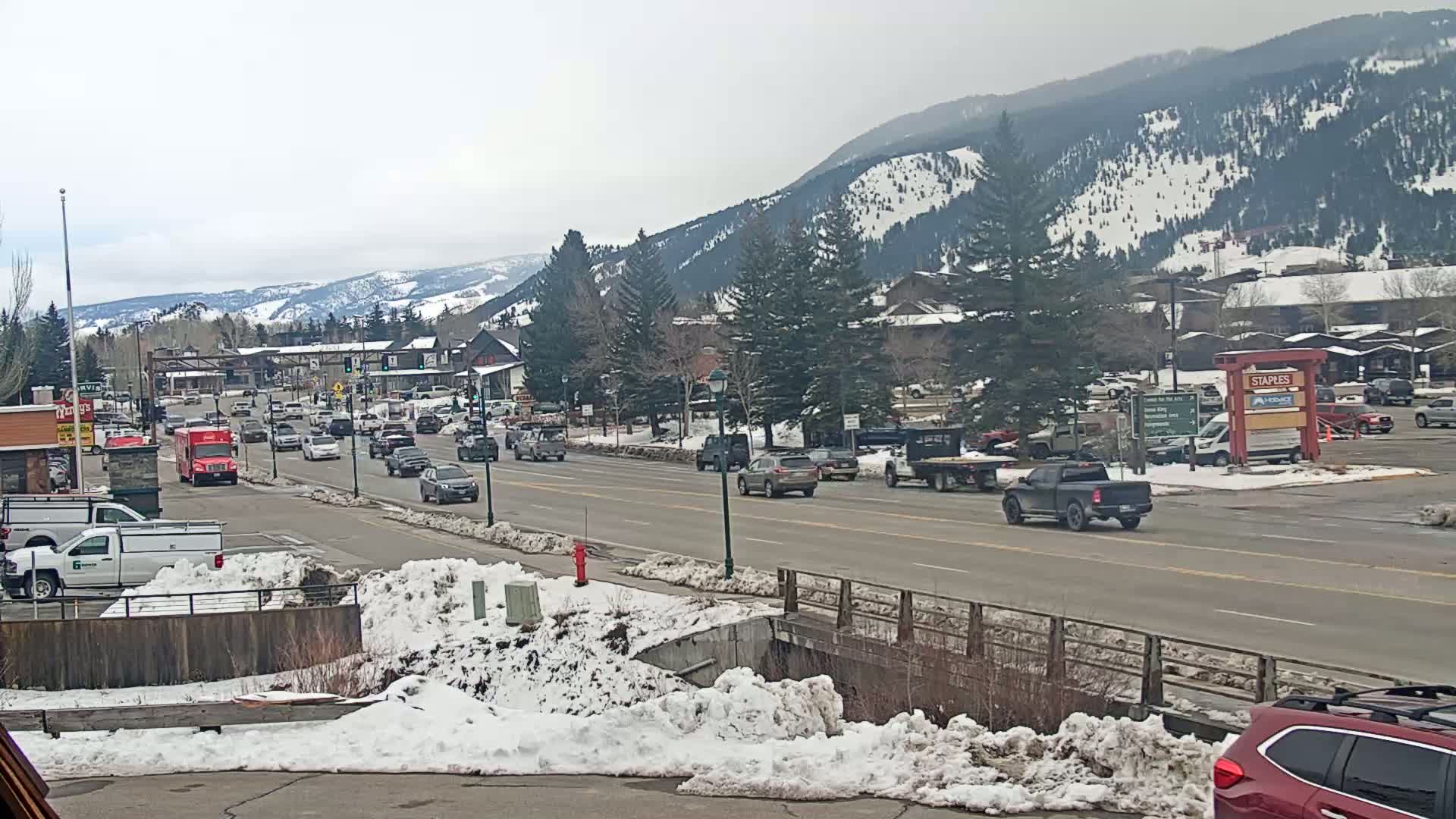 A partly cloudy day reveals a mountainside partially covered in snow, overlooking a busy road with cars and trucks in a town with snow piled along the sides of the road.