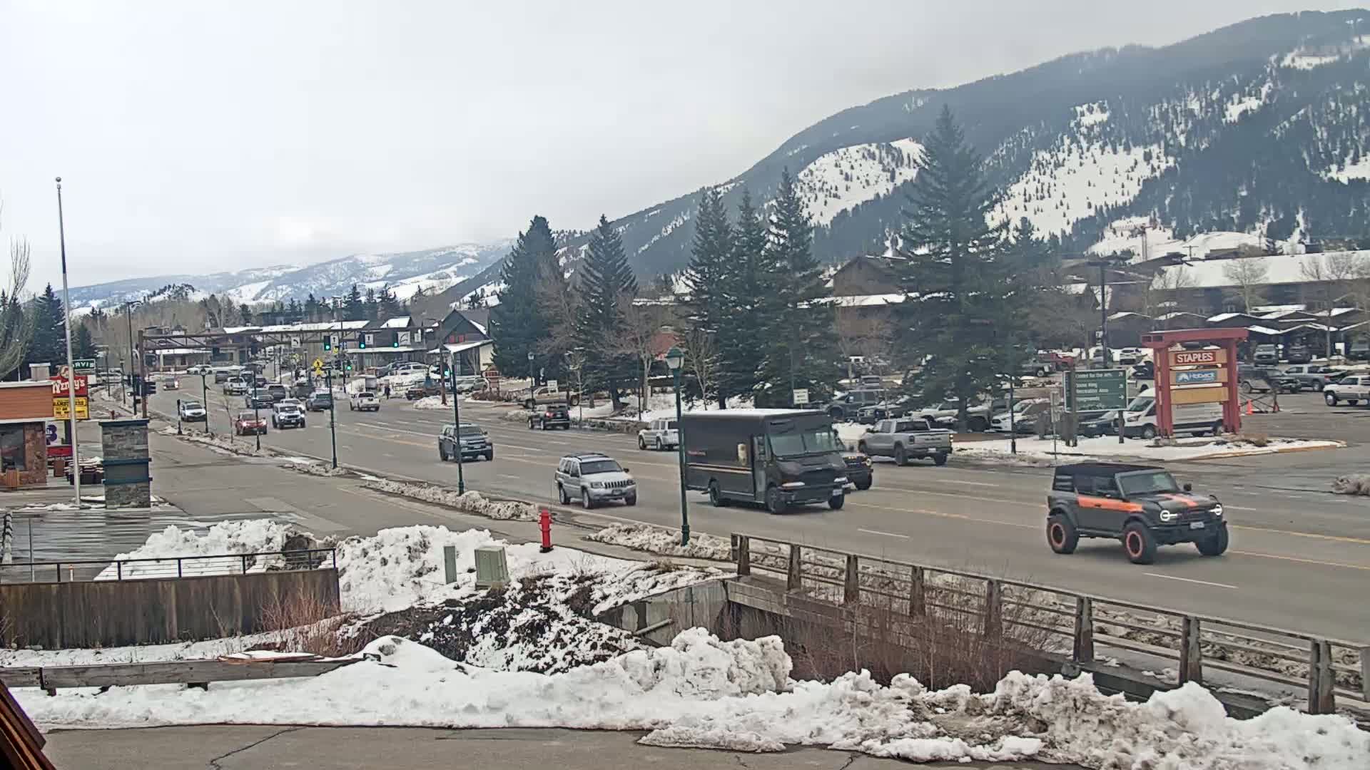A snow-dusted mountain town street scene shows several vehicles driving on a roadway, with snow banks lining the sides of the road and snow-covered mountains in the background under an overcast sky.