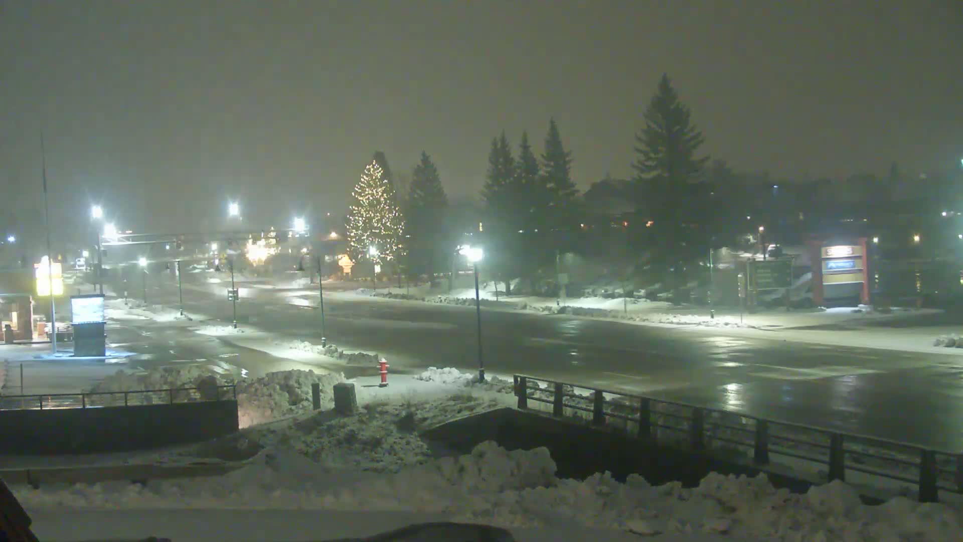 A snow-covered street scene at night, illuminated by streetlights, shows a sparsely trafficked road with a few buildings and trees visible in the background.