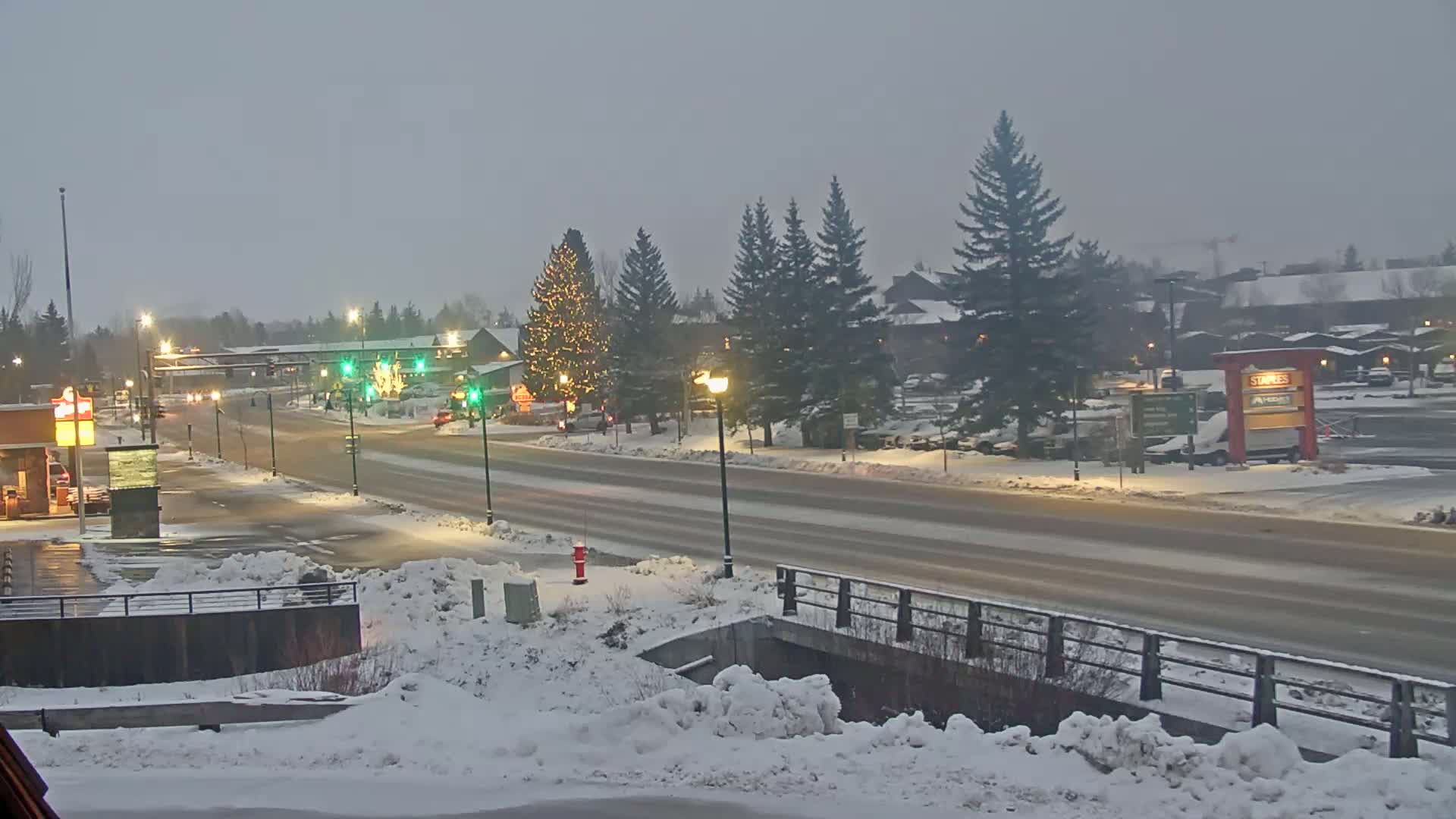 A snow-covered street scene at twilight shows a road with sparse traffic, lined with snow-covered evergreen trees and buildings, some of which are illuminated.