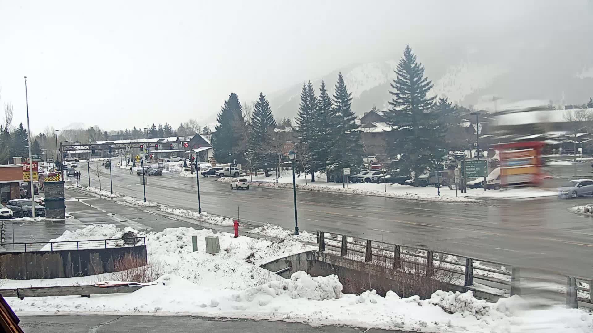 A wet, snowy street scene shows several cars driving past buildings and evergreen trees under an overcast sky, with snow accumulating along the roadside and a mountain visible in the background.