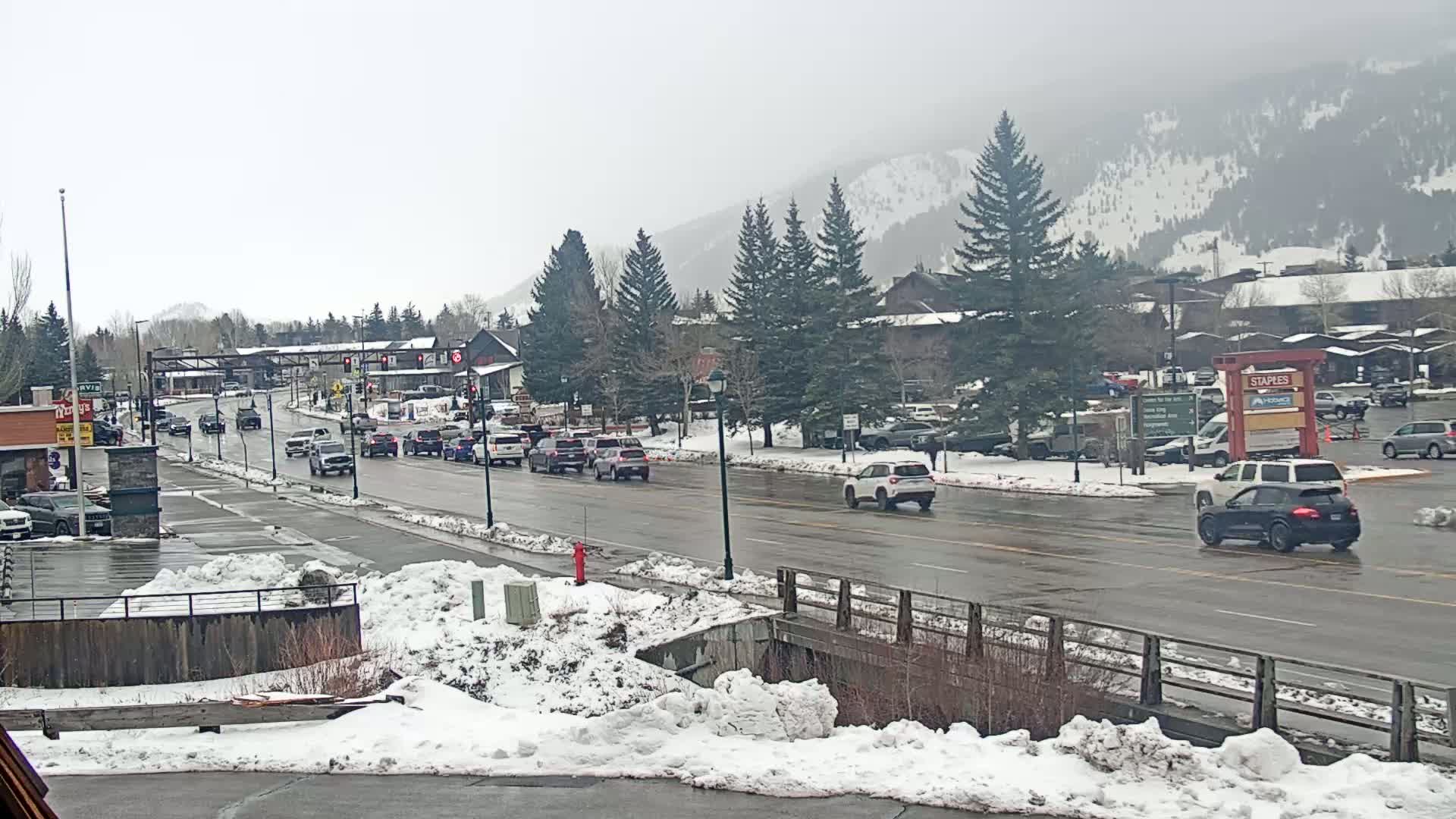 A wet road with moderate traffic flows through a snow-covered town at the base of a snow-dusted mountain range under an overcast sky.