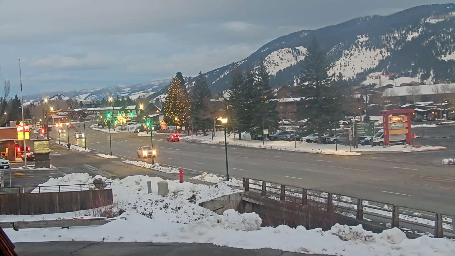 A snow-covered town street scene under a cloudy sky, with cars driving on the road and mountains in the background.