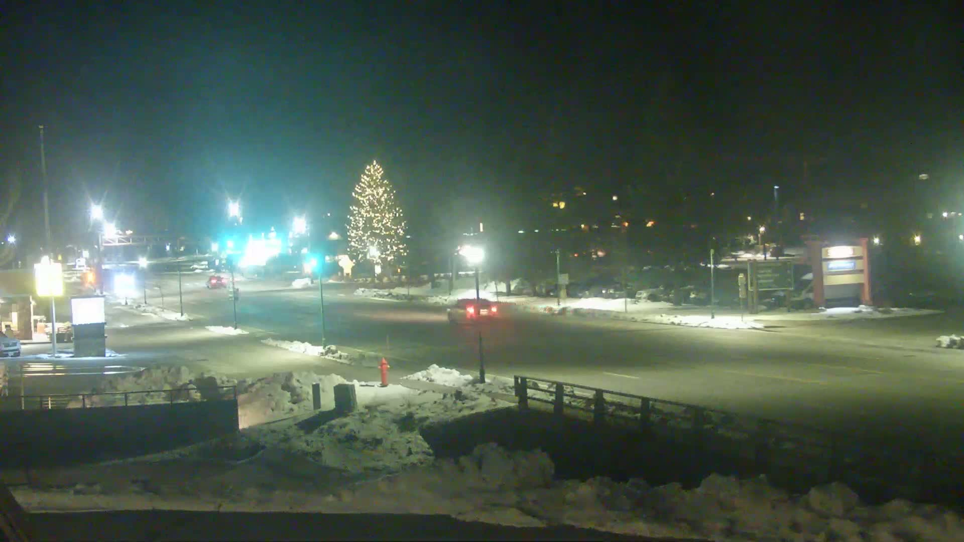 A nighttime scene of a snow-covered street with a few cars and a lit Christmas tree in the distance.