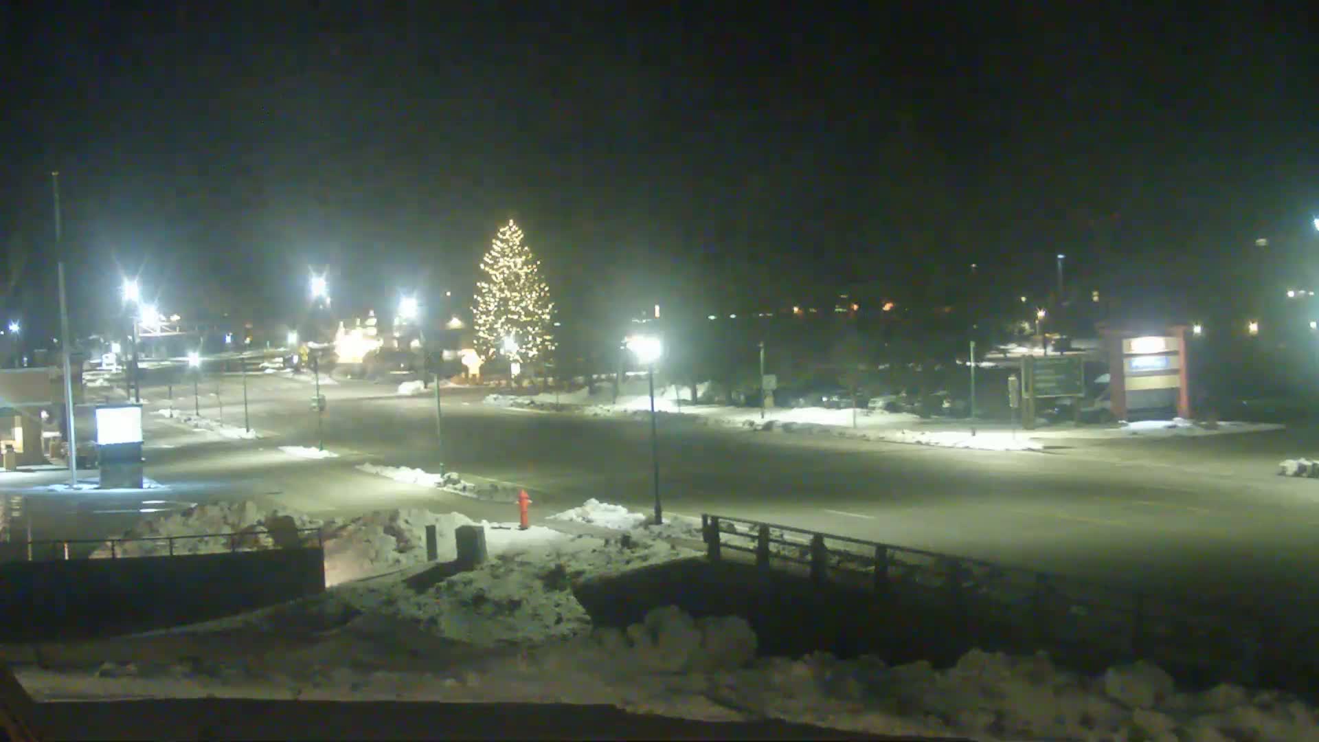 A snow-covered town square at night, illuminated by streetlights, features a large, decorated Christmas tree in the center.