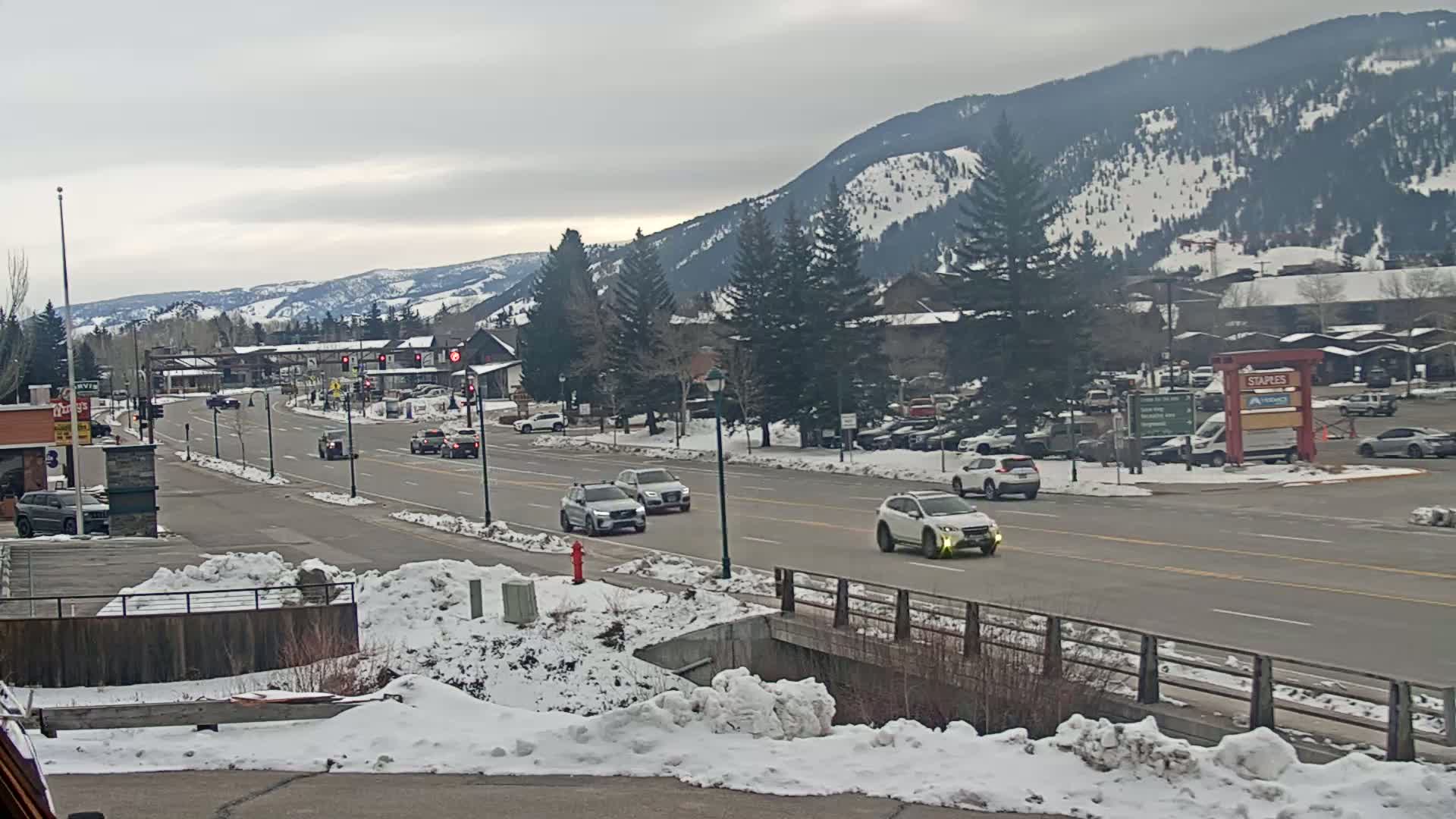 A paved road with several cars driving on it, lined with snow-covered banks and buildings, under an overcast sky and snow-dusted mountains in the background.