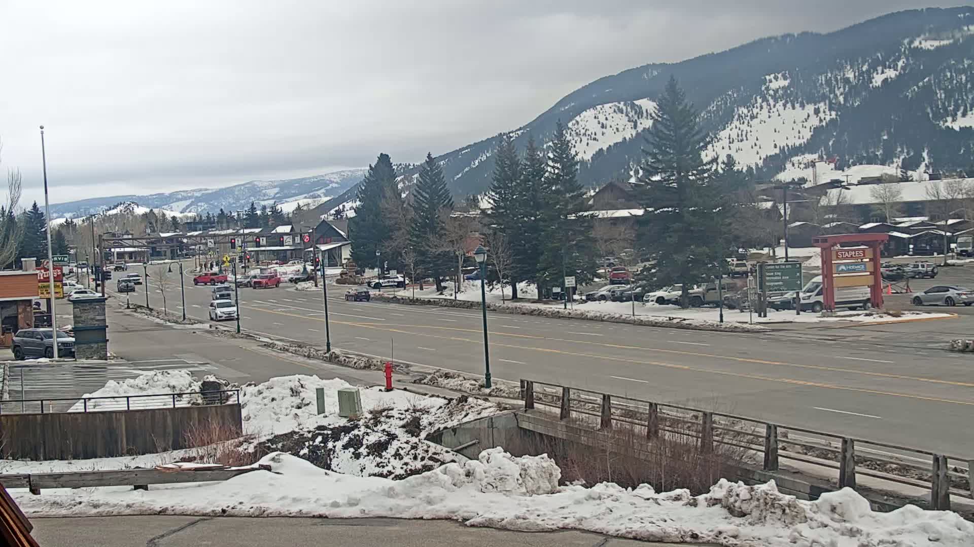 A snow-dusted street scene shows a road with several vehicles driving through a town nestled at the base of snow-capped mountains under an overcast sky.