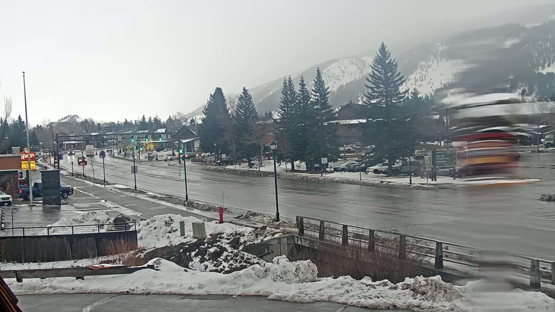 A wet, snow-covered street scene shows a town nestled against snow-dusted mountains under a gray sky, with cars traveling along the road and a blurred vehicle passing by in the foreground.