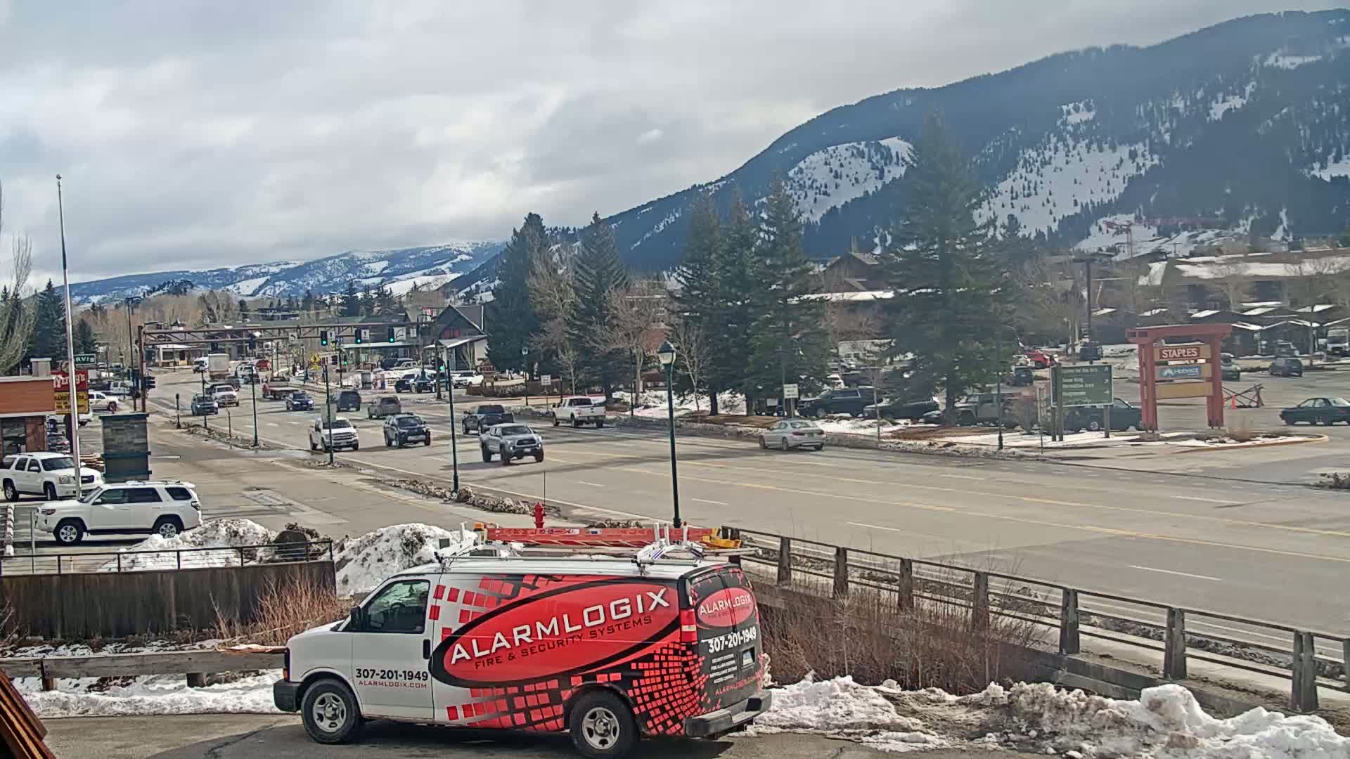 A partly cloudy day shows a mountain-view town with several cars driving on a main road, a security van parked, and snow on the ground in some areas.