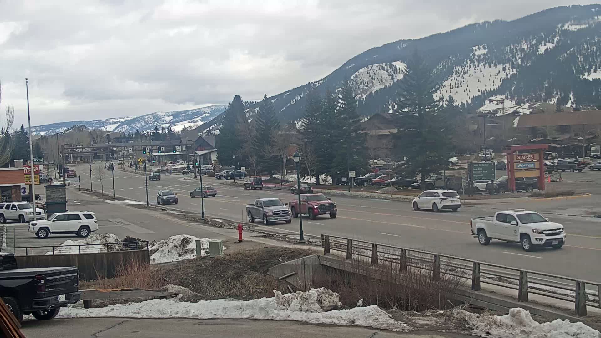 A road scene shows several vehicles driving on a multi-lane road that runs alongside snow-dusted landscaping and a bridge, with snow-capped mountains visible in the background under a cloudy sky.