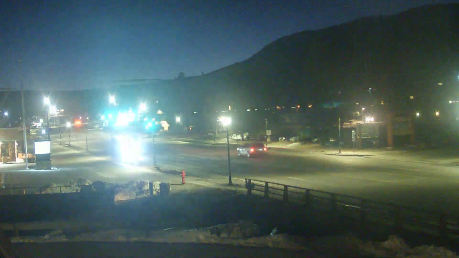 A nighttime view of a sparsely trafficked road intersection, illuminated by streetlights, with a hill in the background and light snow or ice visible on the ground.