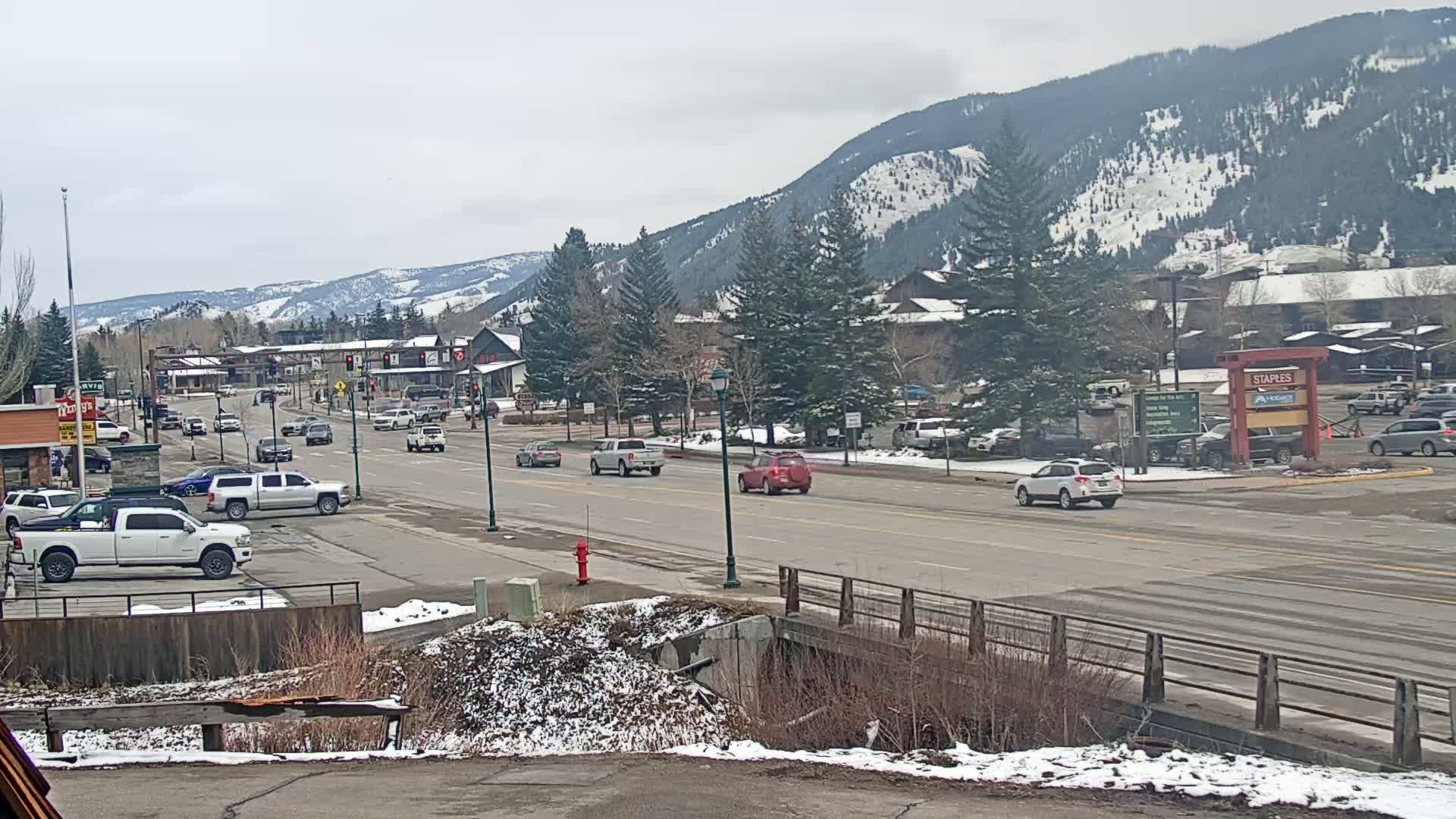 A mostly cloudy day reveals a mountain town street scene with light traffic, snow-dusted ground, and evergreen trees lining the road, all set against a backdrop of snow-capped mountains.