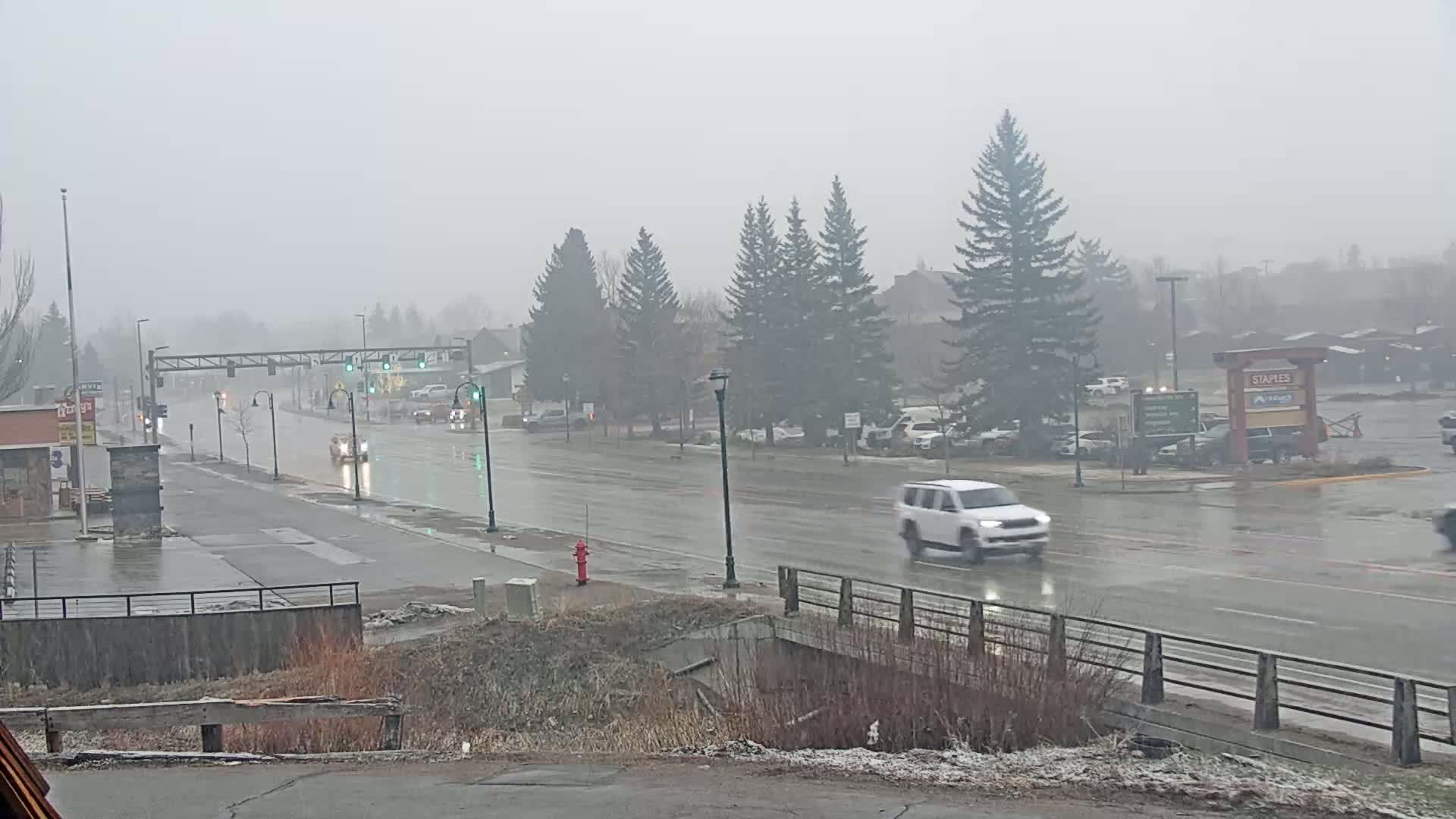 A wet, gray street scene shows several vehicles driving past buildings and evergreen trees in the rain.