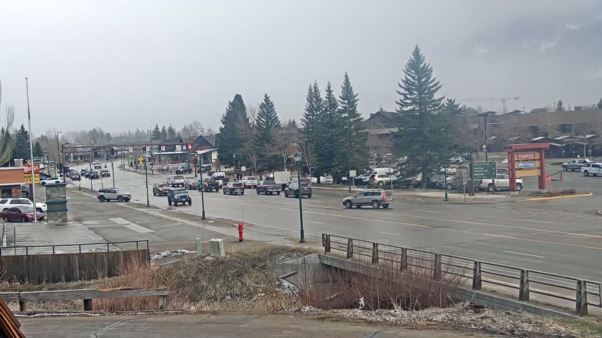 A partly cloudy day reveals a multi-lane road with moderate traffic, lined with buildings and evergreen trees, and a small bridge in the foreground.